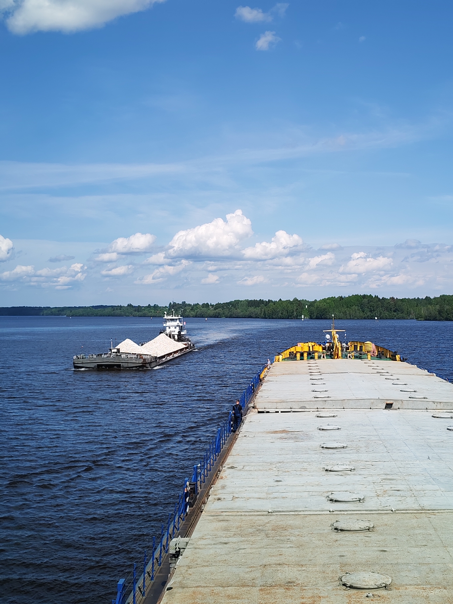 The image shows a wide river in Kondratovo, Russia, under a bright blue sky with scattered white clouds. Two barges are visible on the water. The closest barge is large and grey, with a yellow structure visible towards the back. A smaller tugboat towing a barge filled with what appears to be white sand or gravel is ahead of the larger barge. The water is dark blue with visible ripples. The shoreline on the horizon has dense green trees. A blue railing lines the edge of the large barge, where a few people stand or walk.