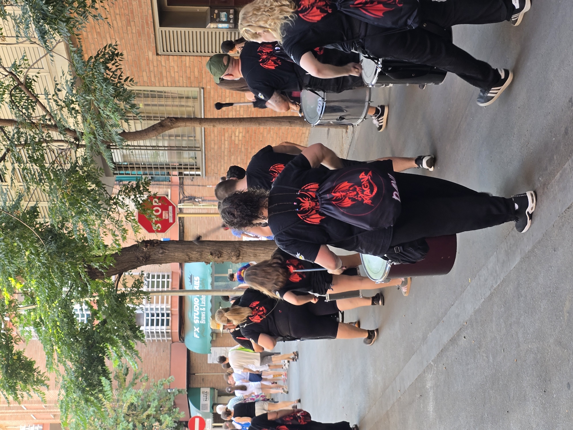 A group of people in black t-shirts with a red dragon design are participating in a parade, possibly in Cornellà de Llobregat, Spain. Several individuals are playing drums, including a large bass drum. The scene appears to be taking place on a street during the daytime, with a building constructed of red brick visible in the background. A tree with green foliage is also present, partially obscuring the building. A red stop sign is visible on a pole, and a green awning with the text "FX STUDIO NAILS Brows & Lashes" is displayed on the building. The people are wearing casual clothing and athletic shoes. The overall atmosphere seems energetic, characteristic of a festive street event.
