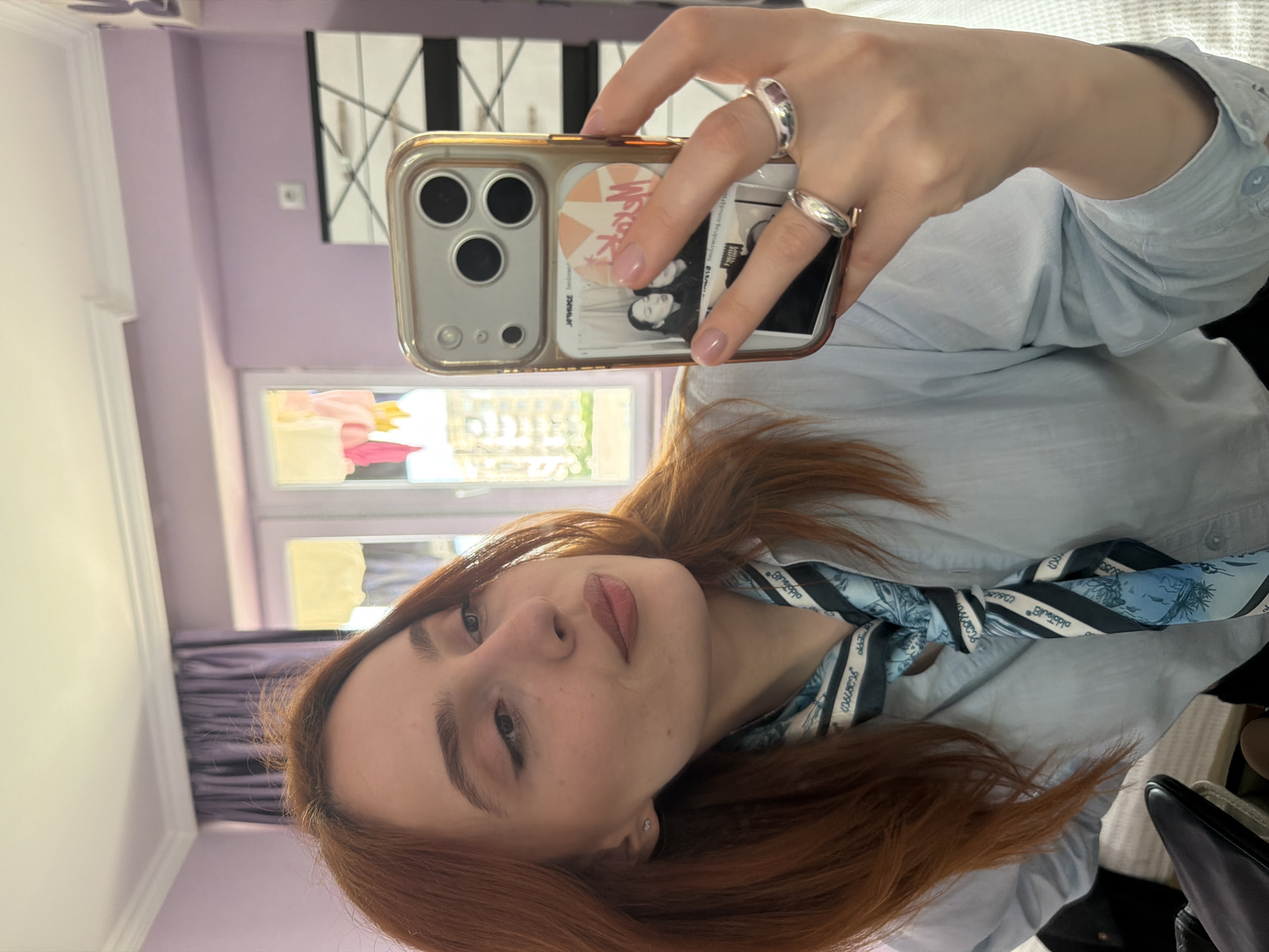 A woman with red hair is taking a selfie. She is wearing a light blue shirt and a blue and black scarf tied around her neck. She is holding a gold iPhone with rings on her fingers. The background includes a window, lavender walls, and a black and white abstract artwork. The specific location in Tbilisi, Georgia cannot be determined from this image.