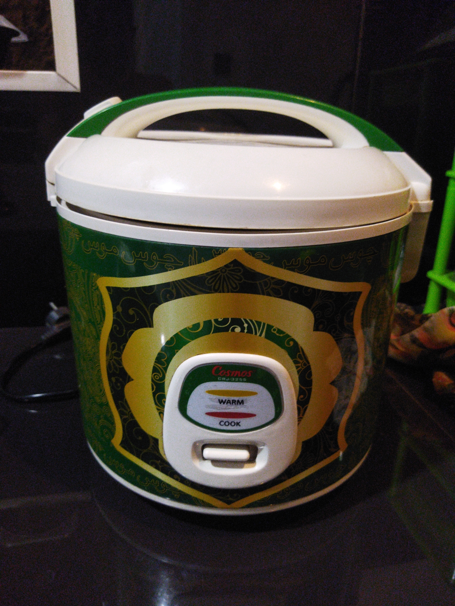 A front-facing, medium shot shows a green and gold rice cooker with white accents. The rice cooker is positioned on a dark, reflective surface, likely a countertop. The rice cooker features a white lid with a green handle and a white control panel. The control panel has three buttons: "WARM," "COOK," and a circular button with the brand name "Cosmos" and the model number "CRJ-3255." The body of the rice cooker is decorated with ornate green and gold patterns. To the left of the rice cooker, a black power cord is visible. In the upper left corner, a portion of a framed picture is visible, suggesting an indoor setting. The background is dark and somewhat blurry, with a hint of green furniture on the right side. The image appears to be taken in Kosambi, Indonesia, given the context provided.