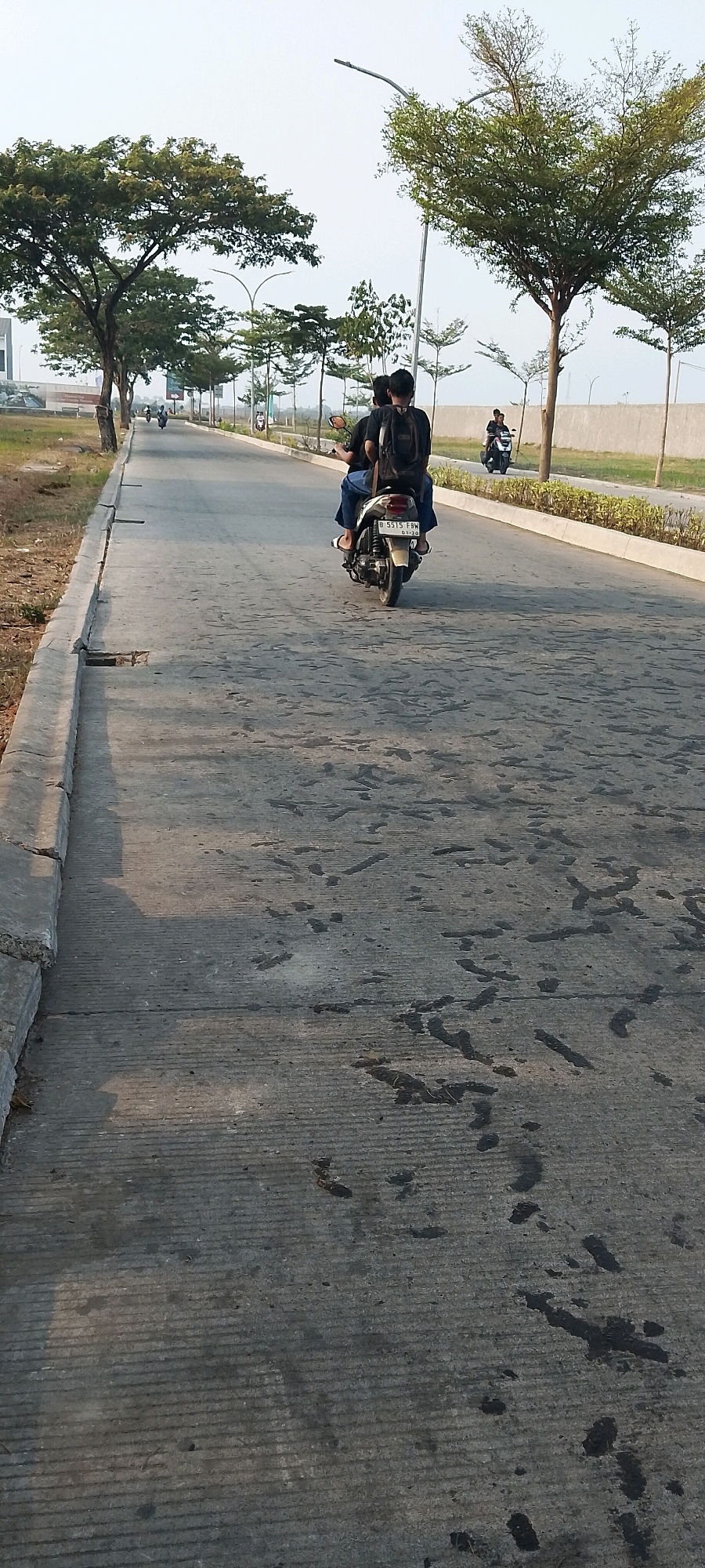 The image depicts a scene on a paved road in Satriamekar, Indonesia, with several trees lining the sides. In the foreground, a concrete curb is visible on the left, with patches of dark, wet-looking stains on the road surface. Two individuals are riding a motorcycle, with the driver in front and a passenger behind wearing a backpack. The motorcycle's license plate reads "B 5515 FBW". In the background, more trees and a grey wall can be seen. Another motorcycle with two riders is further down the road on the right, and a third motorcycle is visible in the distance on the left. The sky is overcast. The overall impression is an outdoor urban or suburban environment.
