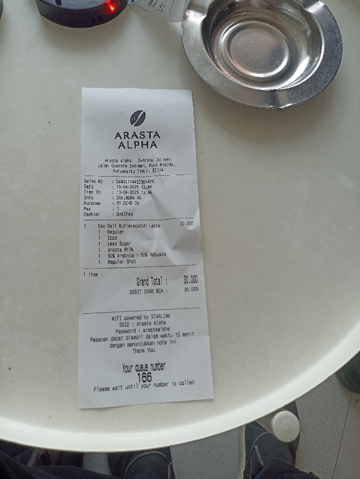 The image shows a receipt for a purchase at Arasta Alpha, a coffee shop located at Jalan Overste Isdiman, Ruko Widoro, Purwokerto Timur, 62114. The receipt details a purchase made on 13-04-2023 at 12:44, with Sales No: SMA01174452500428, Info: DAM.MKRA.45, and Purpose: 01 DIVE JN. The customer, D41FAN, purchased one Sea Salt Butterscotch Latte for 30,000 Indonesian Rupiah. The items included a regular shot of espresso, less sugar, Arasta Milk, 50% Arabica and 50% Robusta. The total cost was 30,000 Indonesian Rupiah, paid for by debit card. The receipt also includes information about the establishment's Wi-Fi and a queue number for the customer's order, 166. The image captures the receipt lying on a white circular table, with a pair of shoes visible at the bottom of the frame. The scene is a cafe in Purwokerto, Indonesia, where a customer has made a purchase and received their receipt.