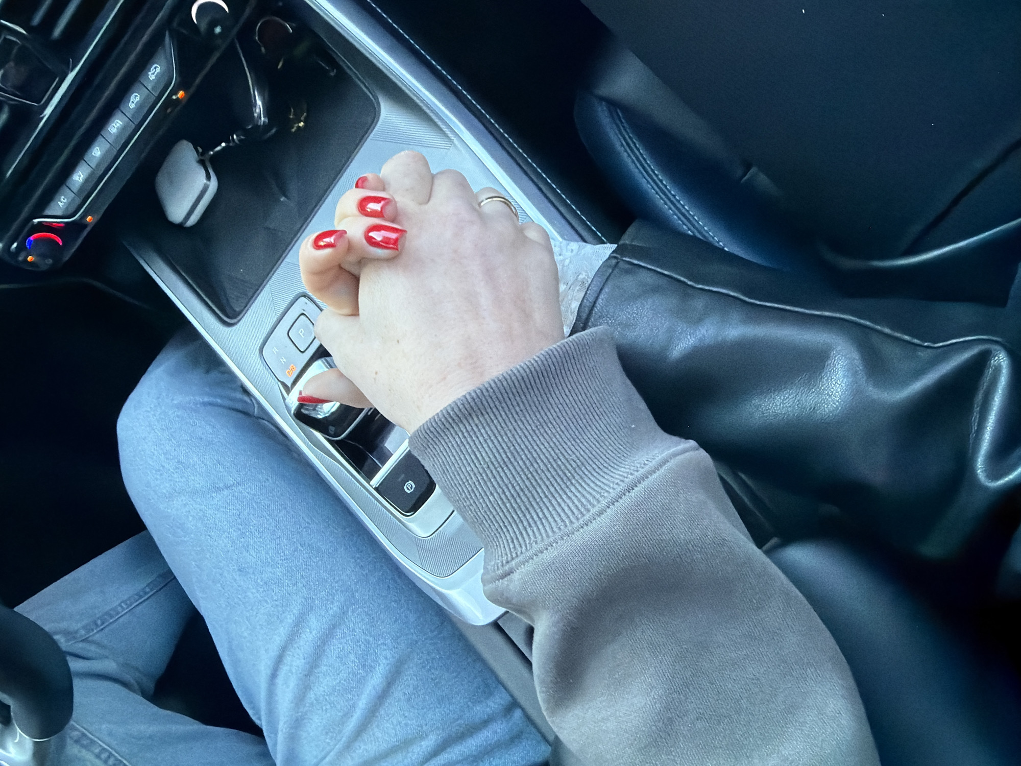 A close-up shot taken inside a car. Two hands are intertwined atop the central console. One hand has red nail polish and a ring on the ring finger. The other hand is partially covered by the sleeve of a gray sweatshirt. The driver wears blue jeans. Visible are parts of the dashboard, gear shift, a car key, and a black leather seat. No location is determined.