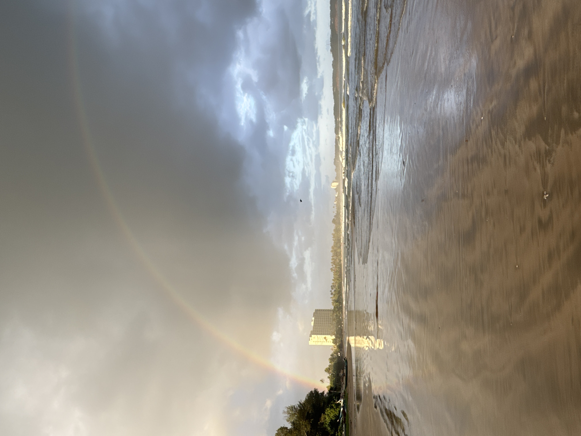 A wide-angle, low-angle shot captures a beach scene with a prominent rainbow arching across a cloudy sky. The foreground is dominated by wet sand, reflecting the sky and creating a mirror-like effect. The ocean is to the right, with gentle waves lapping at the shore. In the distance, a line of palm trees separates the beach from what appears to be a small town or resort. A tall, light-colored building stands out amidst the trees, its reflection visible on the wet sand. The sky is overcast with dramatic clouds, but patches of brighter light suggest either a break in the clouds or the sun is low on the horizon. A faint rainbow is visible on the left side of the frame, a subtle arc of color against the muted sky. The overall mood is serene and slightly dramatic due to the weather conditions. The time of day is likely late afternoon or early morning, given the soft light and the presence of a rainbow after or during rain. The location appears to be a tropical beach, with the building and vegetation suggesting a coastal resort area. There is no visible text in the image.