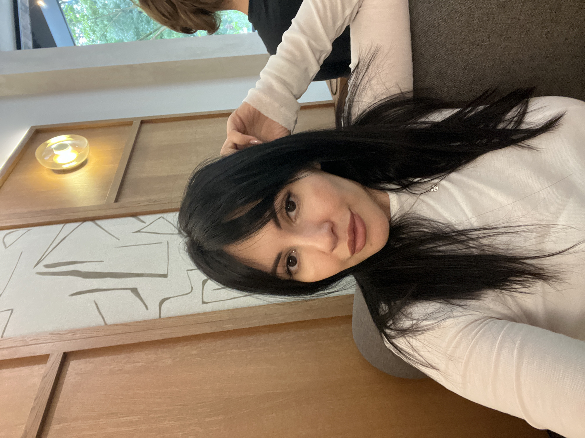 A medium shot depicts a woman with dark hair and fair skin, wearing a long-sleeved white shirt. She appears to be reclining on a brown surface or piece of furniture, possibly a couch or chair. To her left, a wooden wall with geometric patterns is visible, and above, a light fixture emits a warm glow. Behind the woman, another person is partially visible, wearing a black shirt. The image's location is potentially Sochi, Russia, but this is not visually verifiable from the image alone.