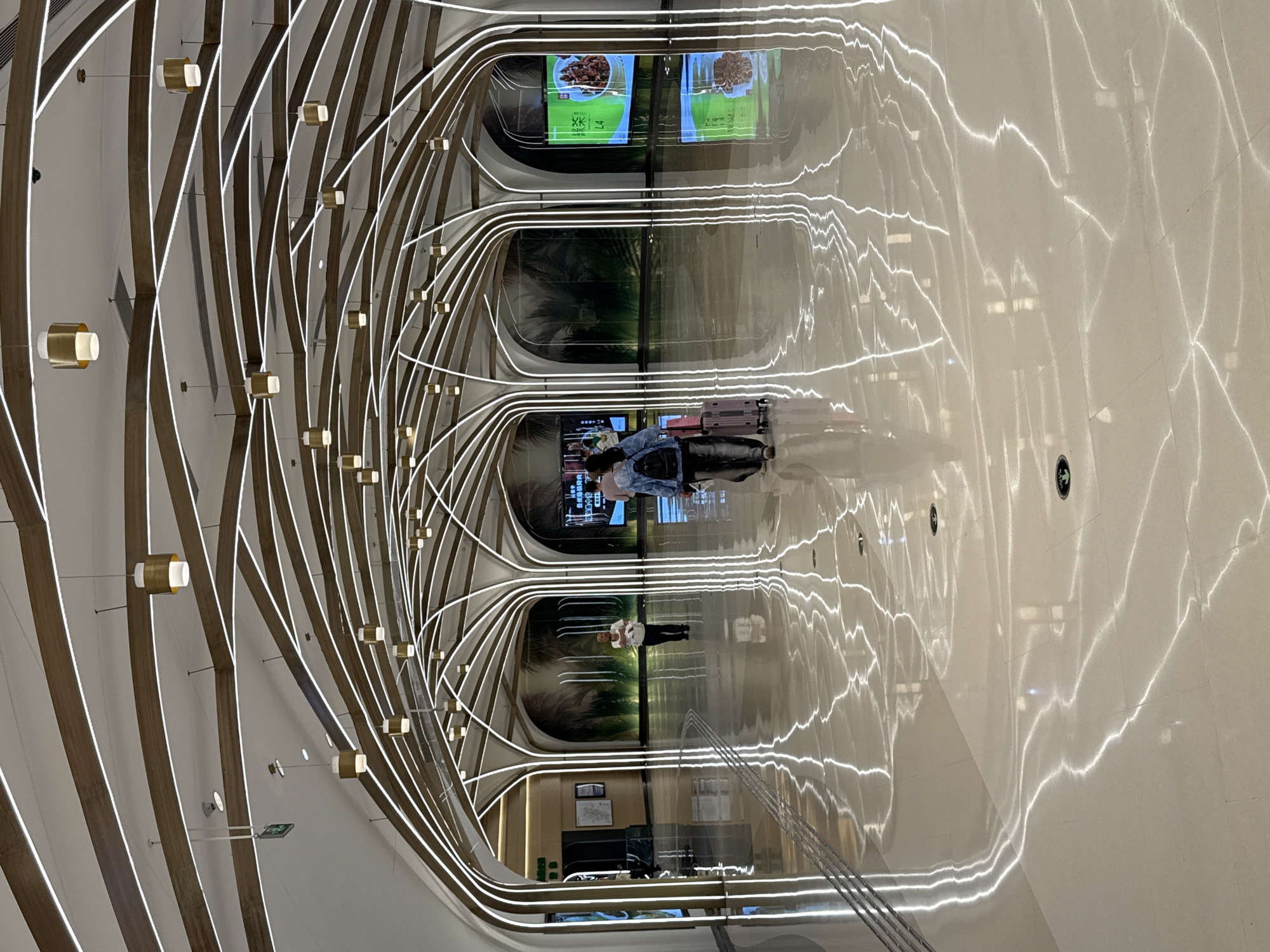 An indoor shot from Liangjiang, China shows an abstract interior. A man and a woman walk, and a woman pulls luggage. Decorative fixtures of wood and neon lights line the ceiling, reflecting in the high-gloss floor. Video screens display food and nature scenes. Signage appears on the wall.