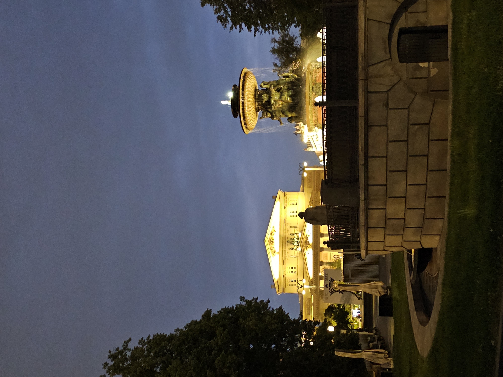 The image depicts a nighttime scene in Moscow, Russia, featuring the Bolshoi Theatre in the background, illuminated by warm yellow lights. In the foreground, a tiered fountain with sculpted figures is active, with water cascading down. A statue of a man in silhouette stands on a pedestal near the fountain, looking towards the theatre. To the left of the theatre, a large tree with dark green foliage is visible. The sky is a deep, dusky blue, suggesting twilight or early evening. The overall atmosphere is serene and stately, highlighting iconic Moscow landmarks.