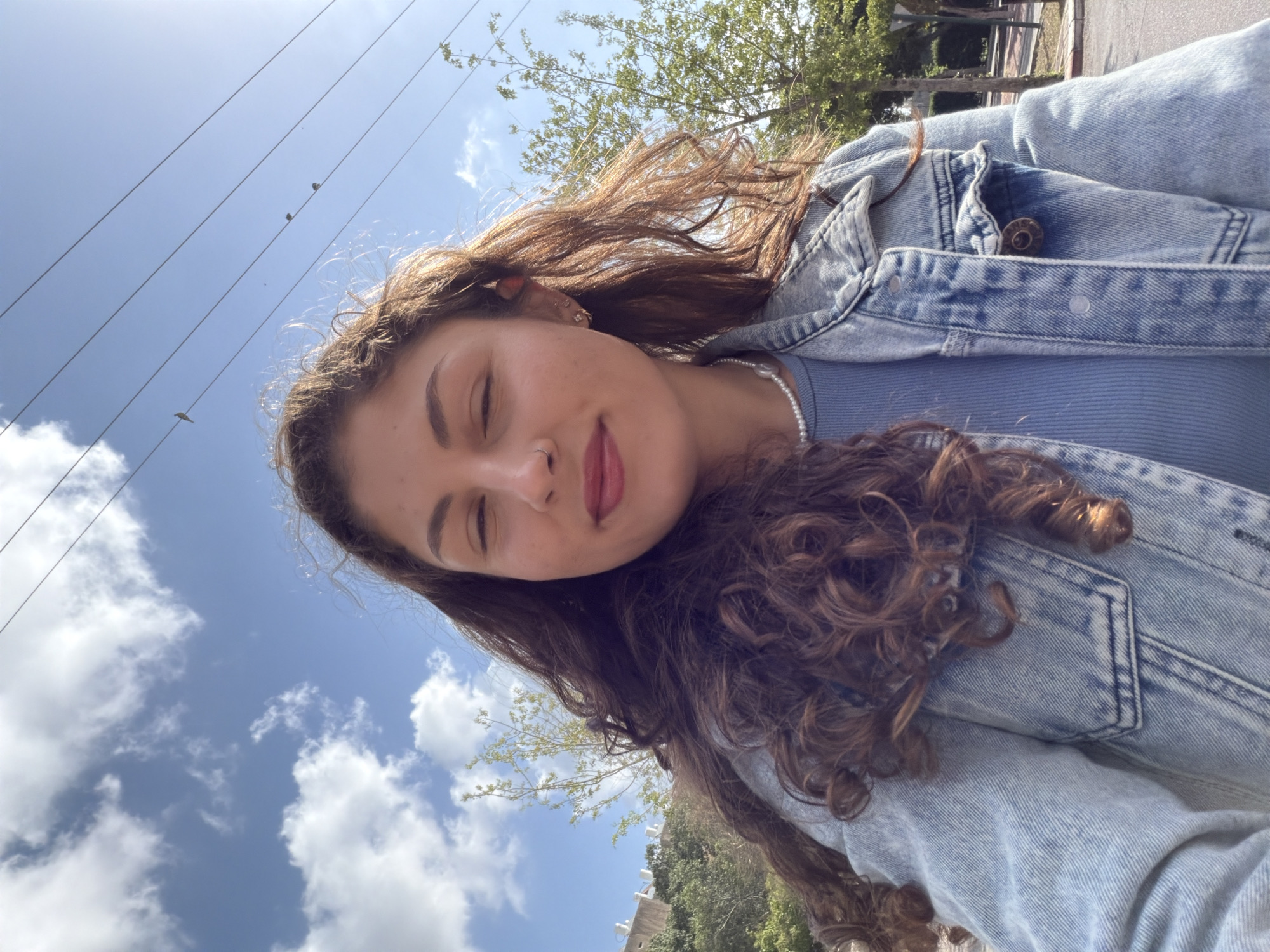 A selfie image shows a young woman wearing a light blue ribbed shirt and a denim jacket. She has long, curly brown hair, and her eyes are closed. She is wearing a pearl necklace and has a nose ring. The background shows a blue sky with white clouds and utility lines with birds perched on them. There are also trees visible, with some greenery and buildings in the distance. The location is likely an outdoor urban area in Kiryat Bialik, Israel.