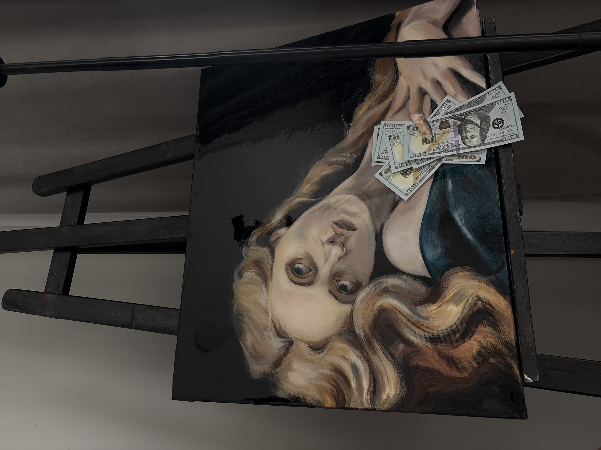 A painting rests on a black easel. The painting is of a woman with long blonde hair, wearing a low-cut dress, holding a stack of US hundred-dollar bills. The background of the painting is black. The easel is set against a light gray wall.
