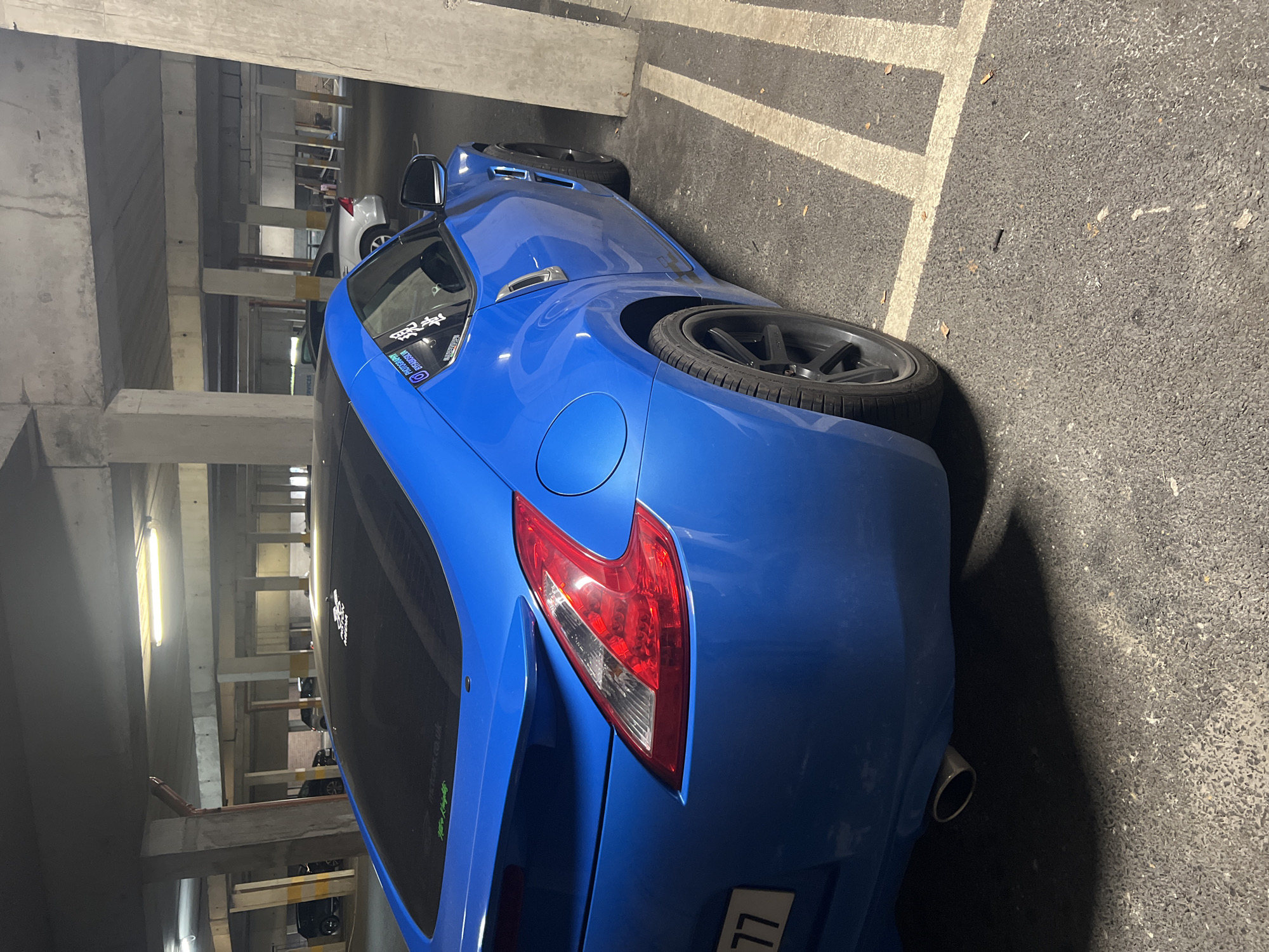 The image shows a blue Nissan 370Z parked in a parking garage. The car is parked in a parking spot marked with white lines. The car's rear is facing the camera, and the license plate is visible. The parking garage is made of concrete and has fluorescent lighting. There are other cars parked in the background.