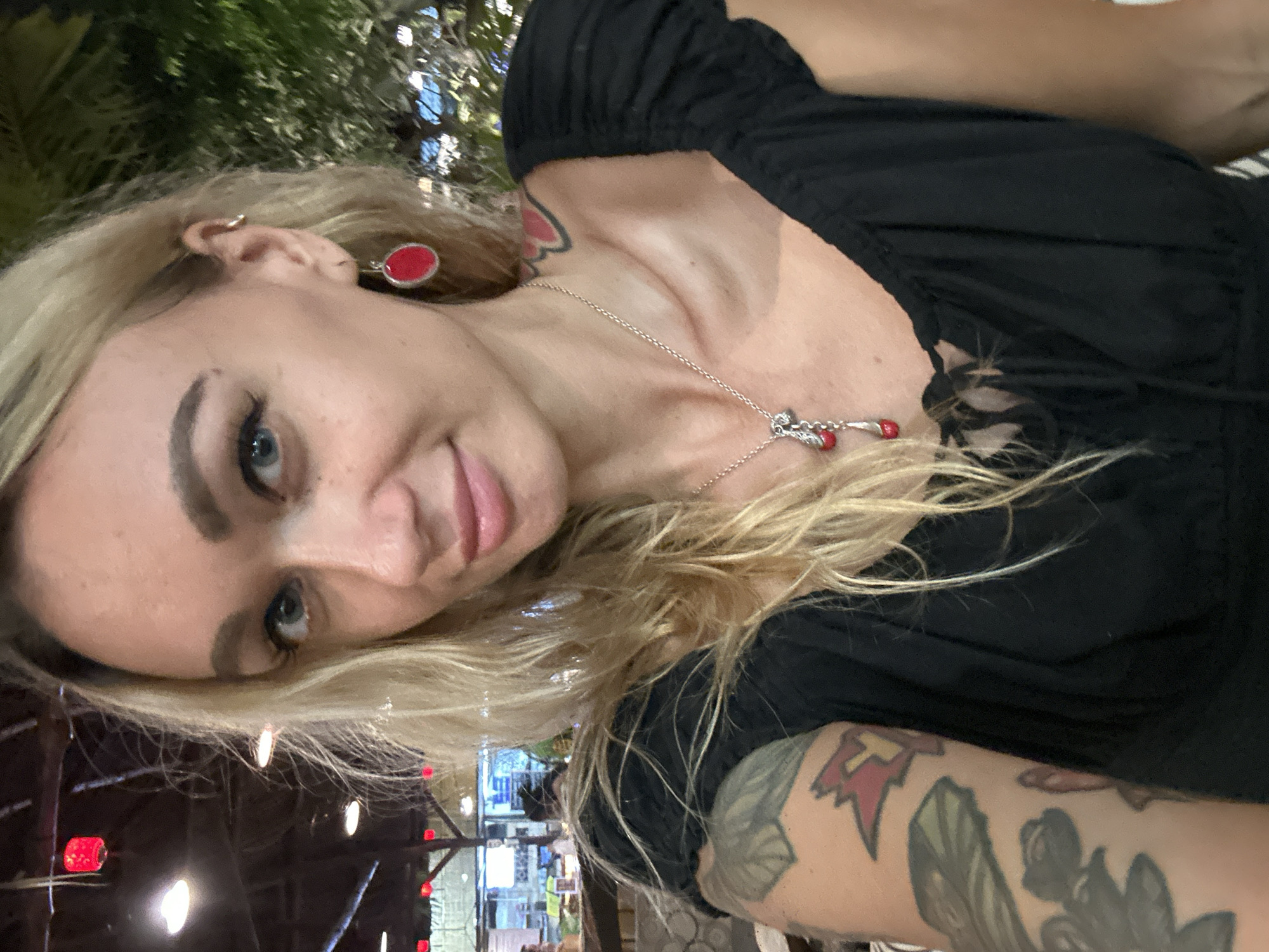 The image is a close-up selfie of a young woman with long blonde hair, light blue eyes, and a slight, pleasant smile as she looks directly at the camera. Her head is tilted, and the image is vertically oriented.
She is wearing a black top with ruffled or gathered detailing on the shoulders. A silver chain necklace with small red beads and a central charm hangs around her neck. She has a distinctive red oval earring in her visible left ear and a small silver hoop piercing in her right ear cartilage. Notable body art includes a red, heart-shaped tattoo on her left shoulder/neck area and a colorful sleeve tattoo on her right arm, which features elements resembling leaves, flowers, and a red, angular symbol.
The setting appears to be an indoor or semi-outdoor establishment, such as a restaurant or bar, given the blurred background filled with soft, warm lights—some red, others white or yellow. Dark green foliage is visible at the top and left of the frame, suggesting the presence of plants or an outdoor patio. The ambient lighting indicates it is evening or nighttime. No text is visible in the image. The location is Phu Thuy Ward, Vietnam.