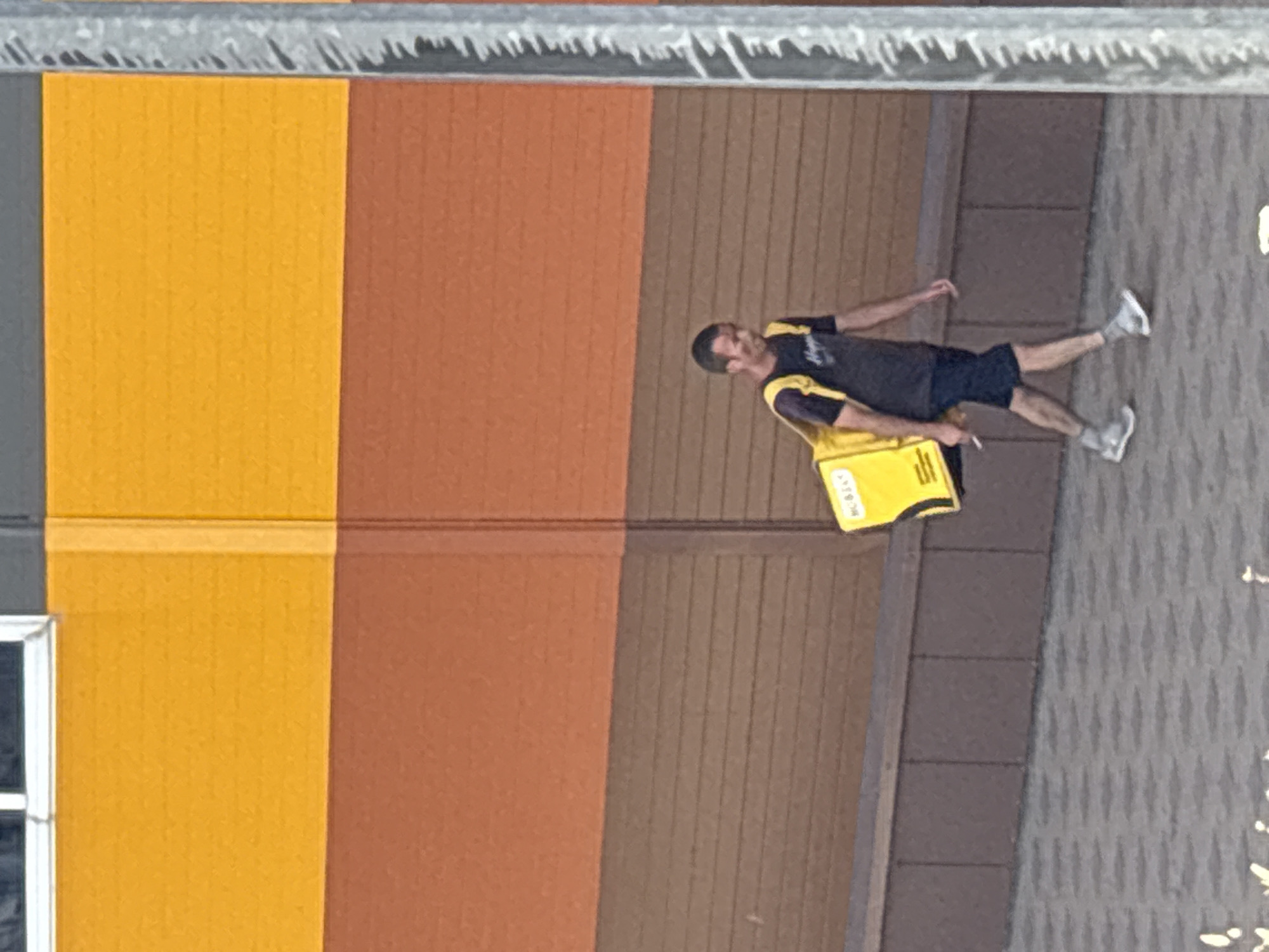 In Rostov-on-Don, Russia, a man walks from left to right across a paved area in front of a building. He wears a dark t-shirt, dark shorts, and grey athletic shoes with socks. A bright yellow insulated delivery bag with black straps is slung over his right shoulder. He is holding a white object, possibly a phone or a receipt, in his right hand. The building behind him features horizontal stripes of color: yellow at the top, then a band of orange-brown, followed by a section of dark brown wood-like paneling. To the right of the frame, a vertical corrugated metal pole is visible. The paving stones in the foreground are a mosaic of grey and white.