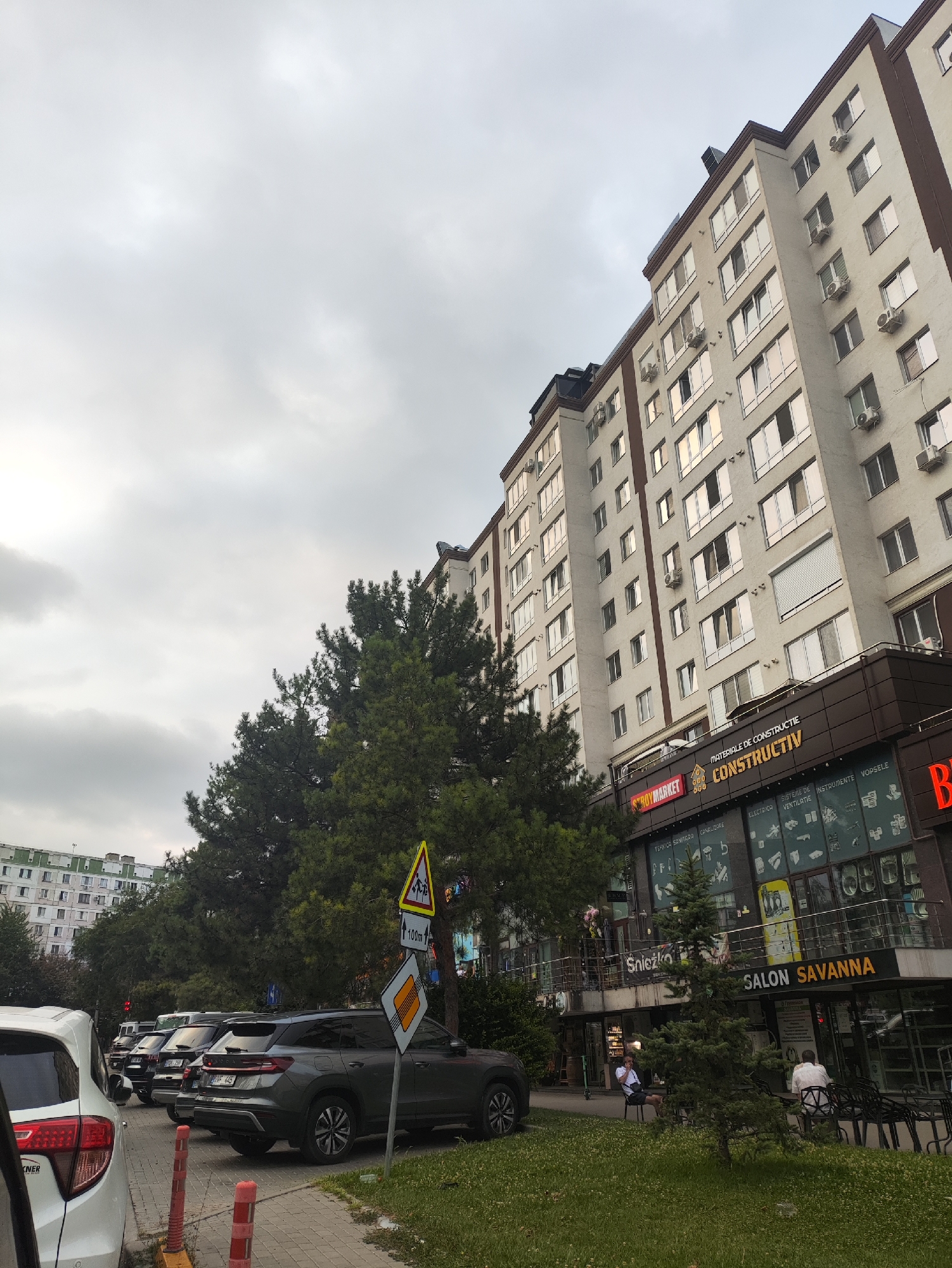 The image captures a view of a modern apartment building in Chișinău, Moldova, under an overcast sky. The building features multiple floors with numerous windows and air conditioning units. At the ground level, there are several commercial establishments, including "STROYMARKET CONSTRUCTIV", "Snježko", and "SALON SAVANNA". A street runs in front of the building, with several cars parked along the side. A few individuals are visible sitting on chairs outside the commercial spaces. Green trees and grass are present in the foreground, adding a touch of nature to the urban landscape. The overall scene depicts a typical day in a bustling city environment.