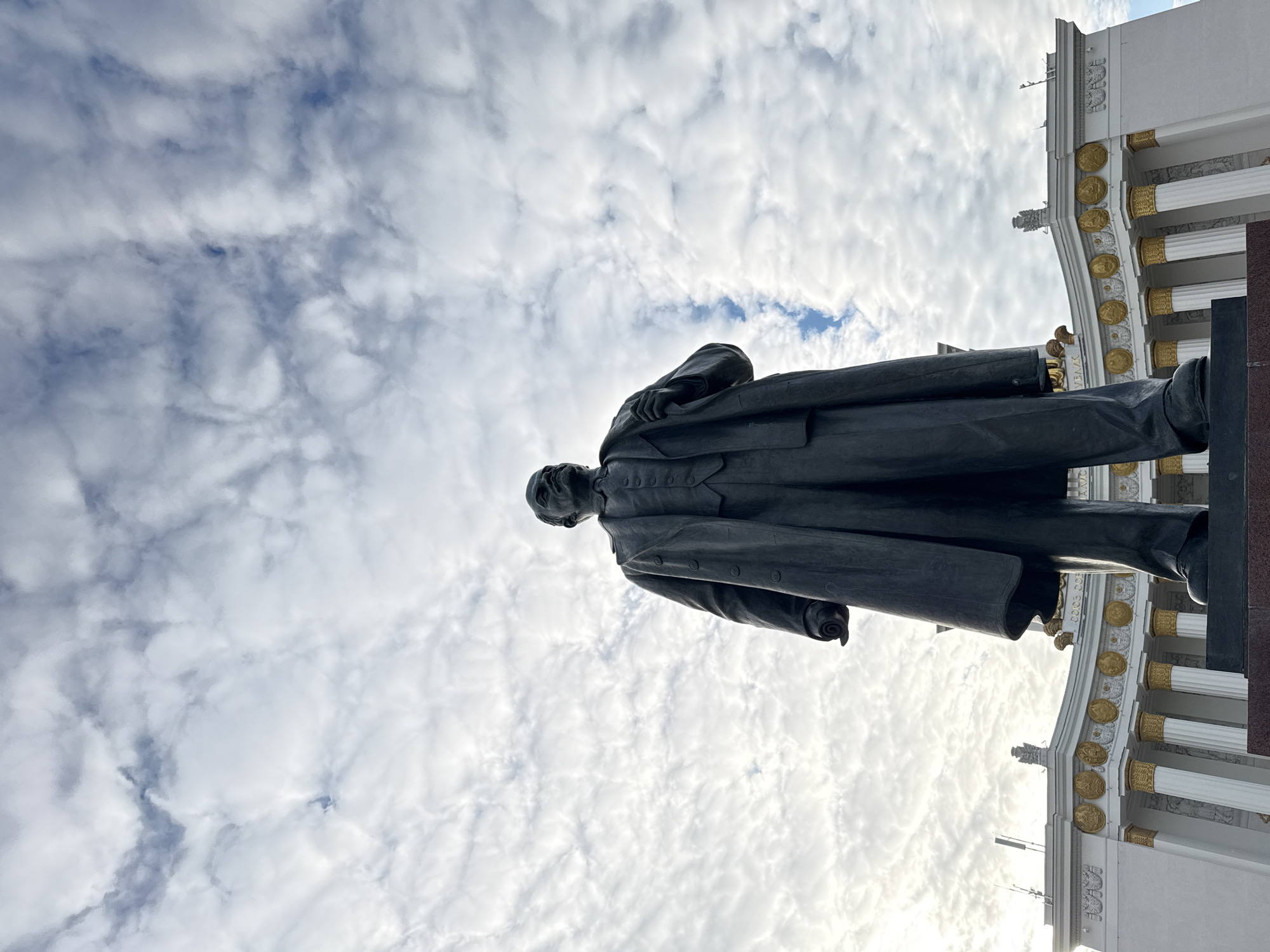 The image captures a low-angle view of a monumental dark statue of a man, set against a partly cloudy sky and a grand, ornate building. The statue, seen from the waist up, depicts a male figure wearing a long, buttoned overcoat and suit, with his left hand gripping the front of his coat. He is looking slightly to the left, and his silhouette is prominent against the brighter sky.
To the right of the statue, a white building with classical architectural features is visible. It features large, fluted columns adorned with gilded capitals and bases. Above the columns, a frieze contains multiple circular, gilded medallions, possibly depicting portraits or symbols, alongside intricate white relief carvings. Partial Cyrillic text, reading "УРАЛ", is discernible on the building's facade below the medallions.
The sky is a mix of white and grey clouds, with some clear blue patches. The lighting suggests daylight, possibly with the sun from the right casting the statue in shadow and illuminating the building's gilded details. The scene conveys a sense of historical importance and architectural grandeur, typical of prominent sites in Moscow, Russia, such as the VDNKh exhibition complex, where the "УРАЛ" (Ural) pavilion is located. No specific activity is depicted; it is a static shot of a monument and its surroundings.