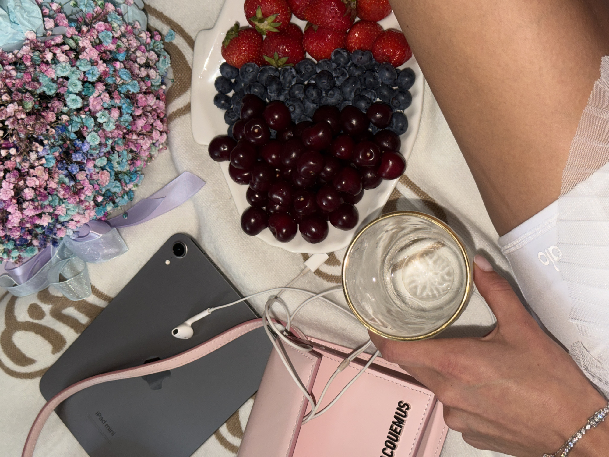 In this top-down, close-up shot, a person's hand holds a clear glass with a metallic rim filled with a clear liquid. The hand, adorned with a diamond bracelet, rests on a white lace-trimmed garment. To the left, a light pink, rectangular clutch with a metallic logo is visible. A dark grey tablet with white earbuds resting on it is positioned above the clutch. Behind the tablet and clutch, a bouquet of small, colorful flowers in shades of pink, blue, and purple is tied with a light purple ribbon. To the right of the flowers and tablet is a white plate filled with a variety of fresh berries: a large pile of dark red cherries, a section of blueberries, and several bright red strawberries. The entire arrangement is laid out on a textured beige surface with a subtle pattern. The scene appears to be indoors, possibly on a bed or sofa, and there is no indication of the location being in Moscow, Russia.