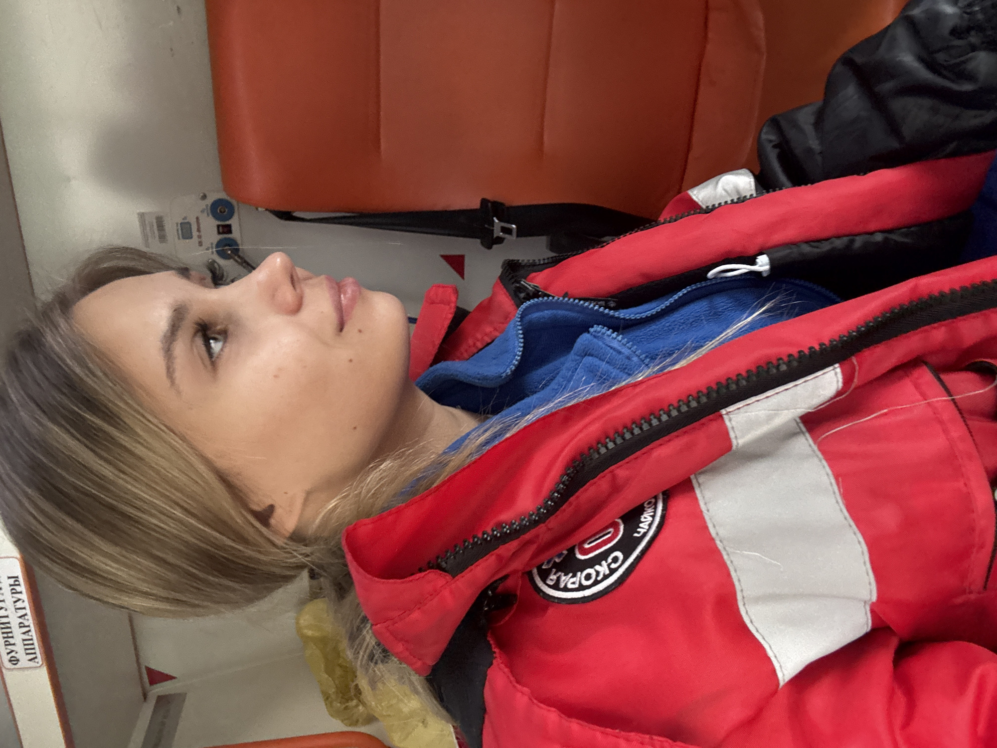 A young, fair-skinned woman with light brown hair lies on an orange surface, possibly a stretcher, inside a vehicle, likely an ambulance. She is wearing a blue fleece jacket under a red paramedic jacket bearing a patch with "Скорая" (Skoraya - ambulance) and "Чайковский" (Chaikovsky) on it, indicating this is likely the city of Chaikovsky, Russia. Her eyes are open and she is looking to the upper left corner of the image. There is a black seatbelt strap visible. Equipment of some sort is mounted on the wall near her head.