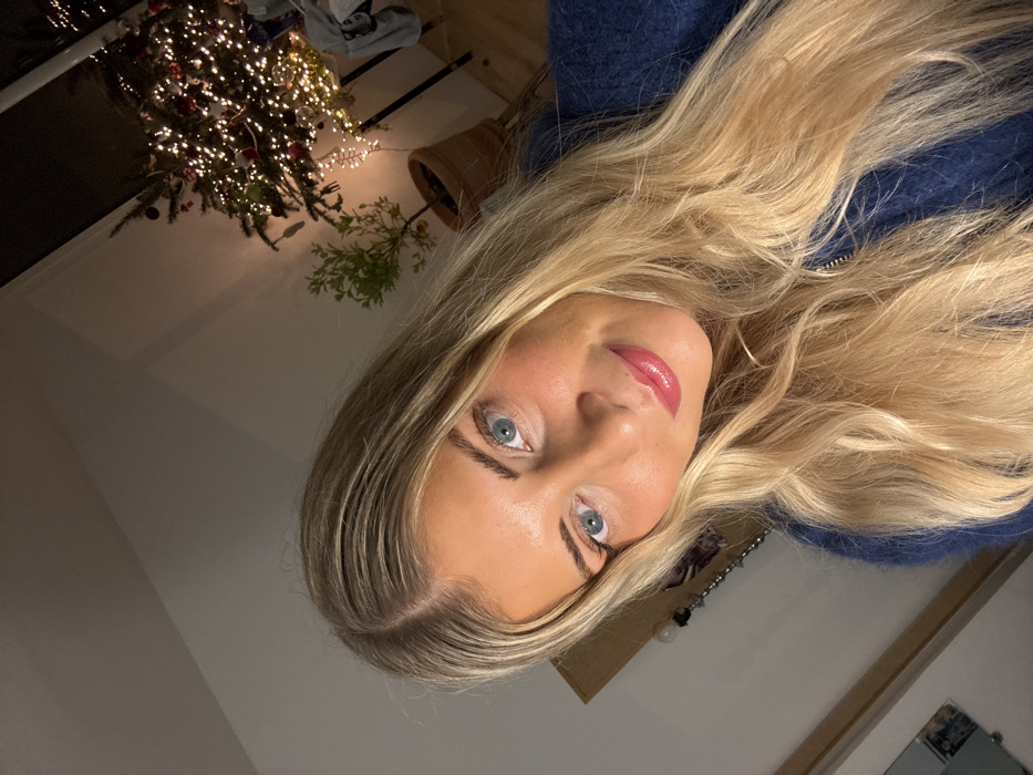 A selfie of a woman with long blonde hair, blue eyes, and pink lips. She wears a blue sweater. In the background, a decorated Christmas tree with warm lights is partially visible. A potted plant also appears in the background, along with a corner of a room with a mirror and wall decoration. The photo is rotated counter-clockwise, creating a tilted perspective.