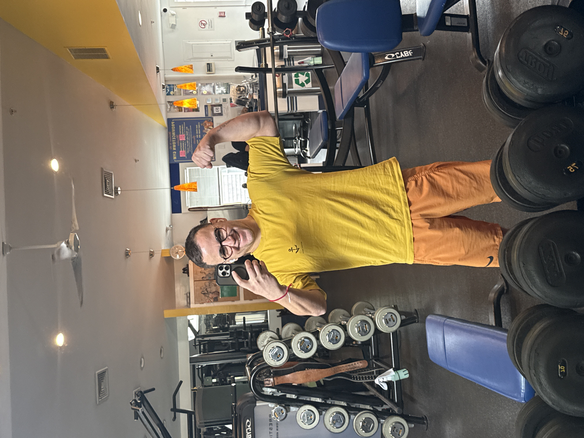 A man in his 40s with short dark hair and glasses is posing for a selfie in a gym. He is wearing a yellow t-shirt with an anchor on it and orange shorts. He is holding his phone in his left hand and flexing his right bicep. He is standing in front of a rack of dumbbells. Behind him is a weight machine with a blue seat. The gym is well-lit with fluorescent lights and has a yellow stripe along the ceiling. The location is Provincetown, United States of America.
