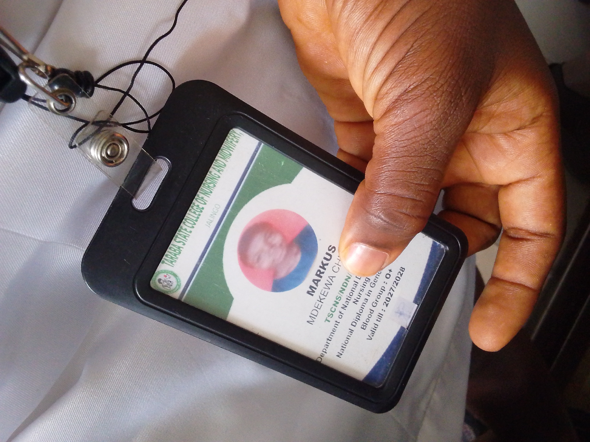 A hand holds a black ID badge holder containing an identification card. The ID card features a circular photo of a person with light-colored hair. Text on the card reads "MARKUS MDEKEWA CH" and lists details such as "TSCNS/NDN/", "Department of National L...", "National Diploma in Gena...", "Nursing", "Blood Group: O+", and "Valid till: 2027/2028". The background of the ID card includes green and white sections with a circular emblem. The ID badge holder is attached to a lanyard with a black cord and a metal clip. The individual's skin tone is dark brown, with visible finger ridges. The surrounding environment appears to be a white fabric surface, possibly clothing or bedding, and a dark background. The overall scene suggests the presentation of an official identification document.