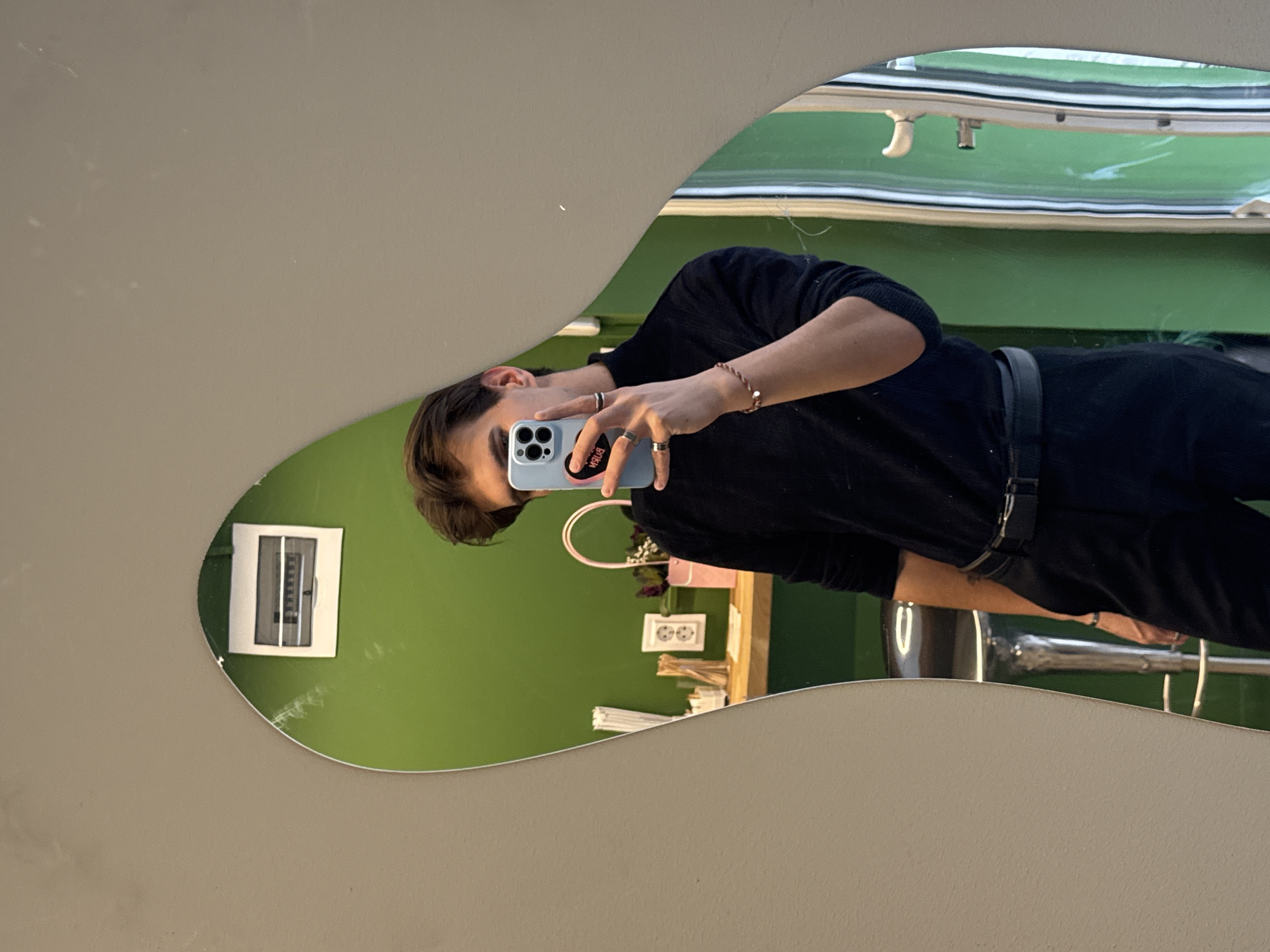 A male individual is observed taking a mirror selfie within the premises of БУРН (BURN) cafe in Sofia, Bulgaria. The subject, visible from the chest to the upper thighs, wears a dark turtleneck, dark trousers, and a black belt with a silver-colored buckle. His left hand holds a light blue smartphone, partially obscuring his face, which features a triple-lens camera array. Multiple rings adorn his fingers, and a braided bracelet with a small silver charm is present on his left wrist. The phone case displays a black sticker with the inverted text "BUЯN". The irregularly shaped mirror reflects a vibrant lime green wall, on which a white electrical circuit breaker panel is mounted in the upper left corner. A wooden counter below the panel holds a white electrical outlet, a clear container of white stirrers or similar slender objects, a small pink bag containing dark flowers or foliage, and a dark green box. A chrome-legged bar stool with a dark seat is partially visible in the reflection to the lower left, and a light-colored door frame or vertical panel appears on the right. The wall surrounding the mirror is a plain beige or light grey.