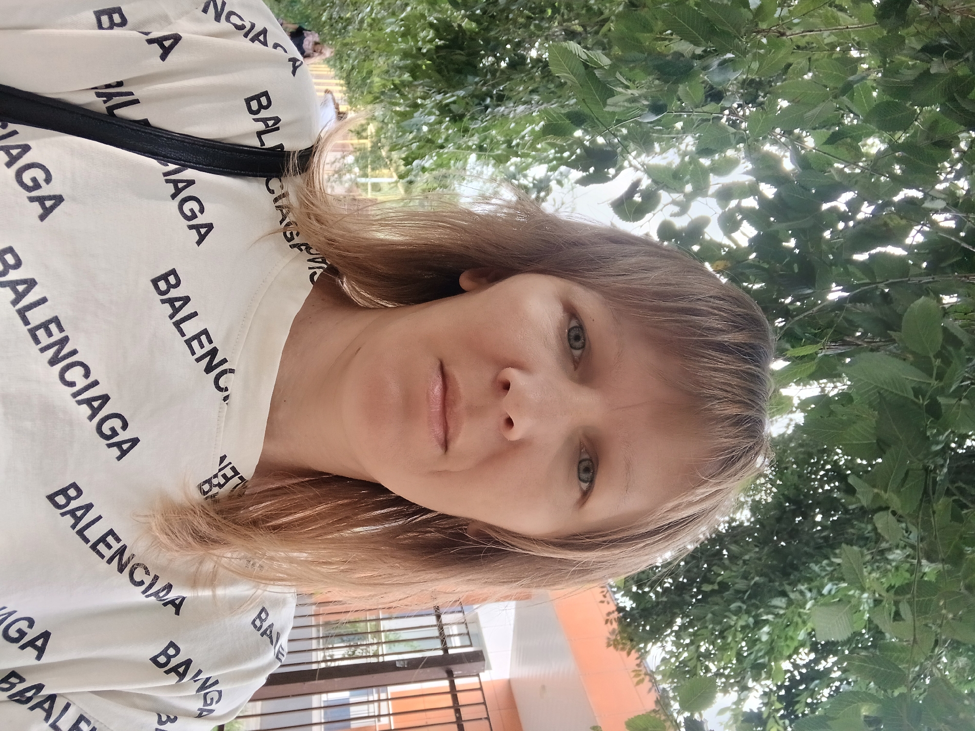 A medium shot captures a fair-skinned woman with shoulder-length light brown hair, facing forward under the shade of green trees. She is wearing a white t-shirt with the word "BALENCIAGA" repeatedly printed in black. The background shows a building with orange and white walls, and a black metal fence. The overall impression is of an outdoor portrait, possibly in a city park or residential area. The image does not provide specific clues to its location in Cherkessk, Russia.