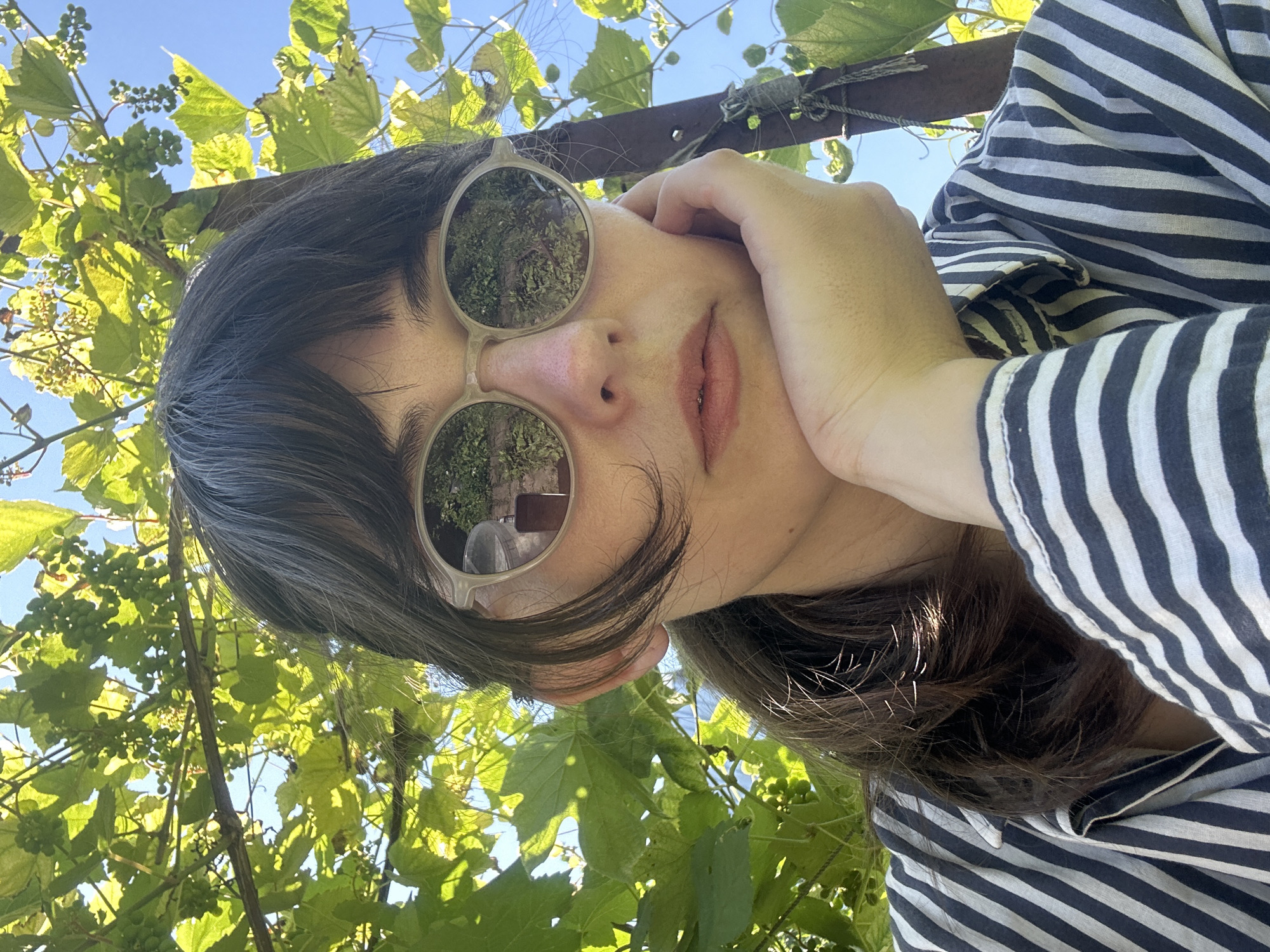 A woman with dark hair and bangs is looking at the camera, wearing sunglasses and a striped shirt. She is resting her chin on her hand. The background consists of green leaves and a blue sky, suggesting an outdoor setting. The image appears to be a selfie. The location is Zvonarevka, Russia.