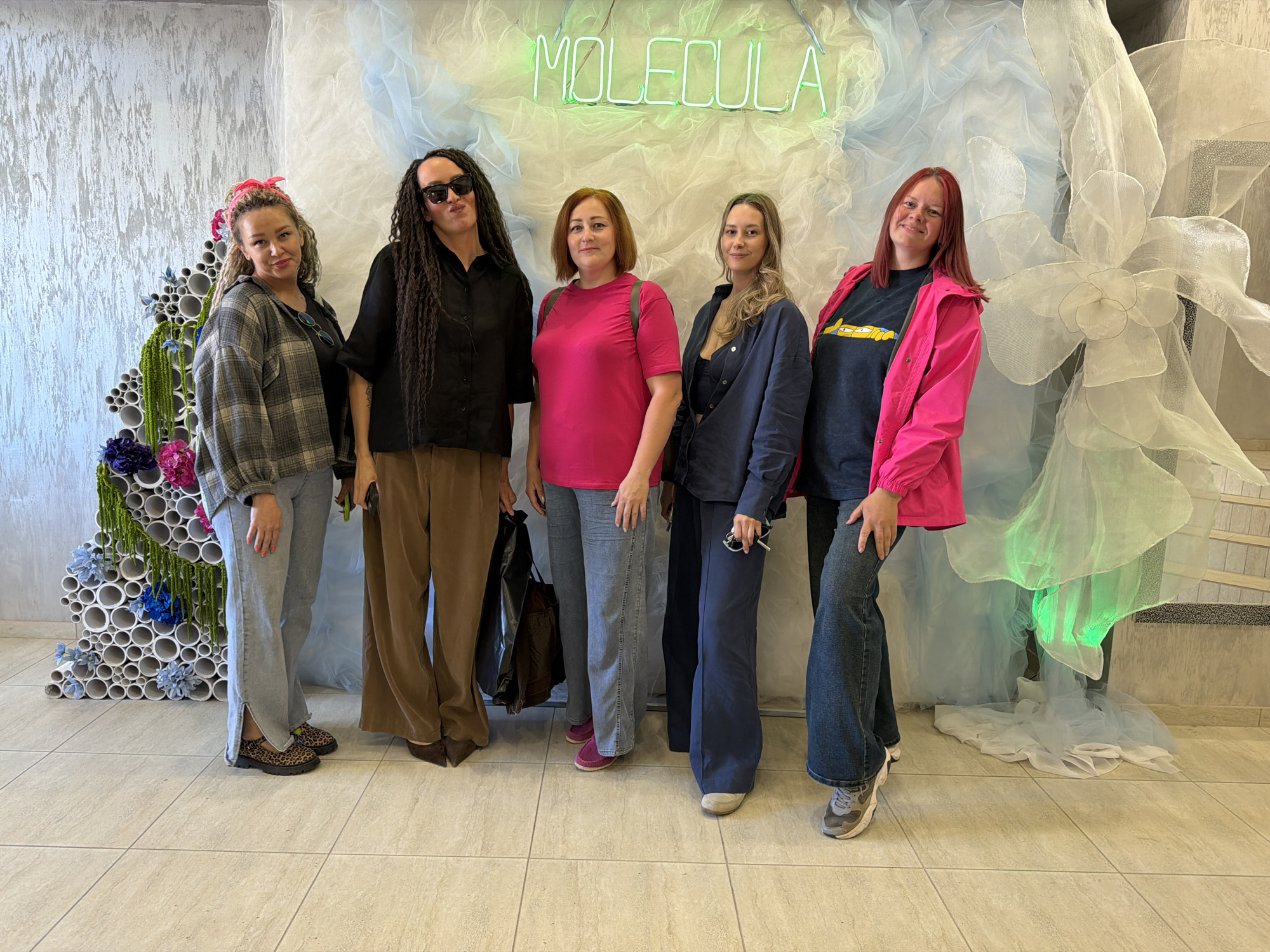 Five women are standing in front of a decorative wall display with a neon sign that reads "MOLECULA." To the left of the women is a sculpture composed of white pipes with blue and purple flowers. The woman on the far left is wearing a gray plaid shirt, light-wash jeans, and animal-print shoes. Next to her is a woman with long dreadlocks, sunglasses, a black blouse, and brown wide-legged pants. The middle woman is wearing a pink t-shirt and blue jeans with pink shoes. The fourth woman is wearing a navy blazer, dark top, and navy pants. The woman on the far right is wearing a dark t-shirt with a cartoon character, jeans, and a bright pink jacket. The background is a decorative wall with white fabric and a neon sign. The floor is tiled. The photo was taken in the city of Chaikovsky, Russia.