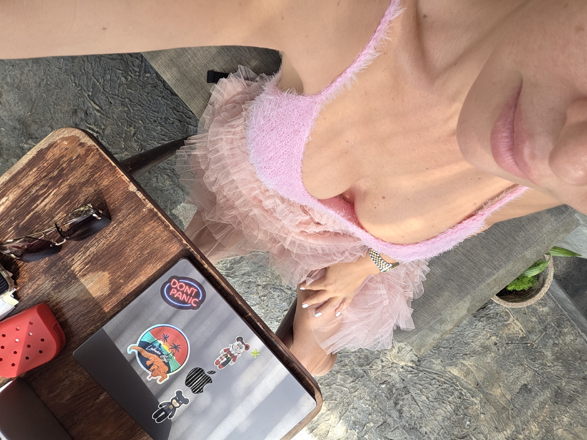 In North Kuta, Indonesia, a woman is seated at a wooden table outdoors, working on a laptop. She is wearing a pink, fluffy-trimmed bralette and a pink tulle skirt. Her hand, with white-painted fingernails, rests on her lap. On the laptop are several stickers, including a Mickey Mouse figure, a "Don't Panic" sticker, and a sticker of a dog with palm trees. To the left of the laptop are sunglasses, and to the right is a red phone case. The ground is a textured, grey stone surface. A small plant in a pot is visible in the background, resting on a grey cushioned chair. Sunlight casts shadows across the scene.