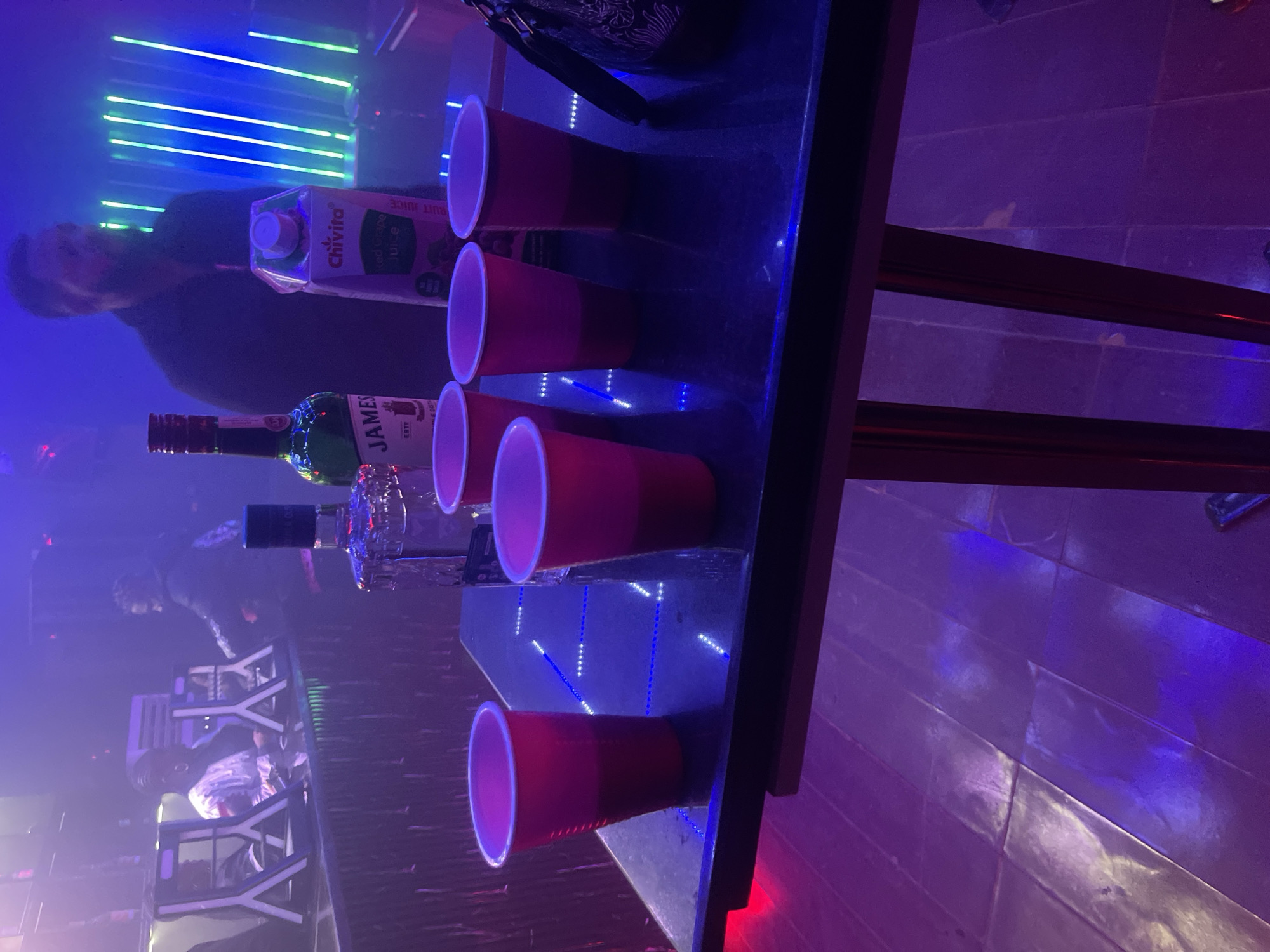 A dimly lit bar in Bar Gada, Nigeria, features a table with several red plastic cups, a bottle of Jameson Irish Whiskey, and a carton of Chivita Red Grape Juice. In the background, a man with a beard, presumably a patron or staff member, is visible near neon lights. The bar's ambiance is enhanced by purple and blue lighting, reflecting on the floor. The overall scene suggests a casual gathering or a night out at the establishment.