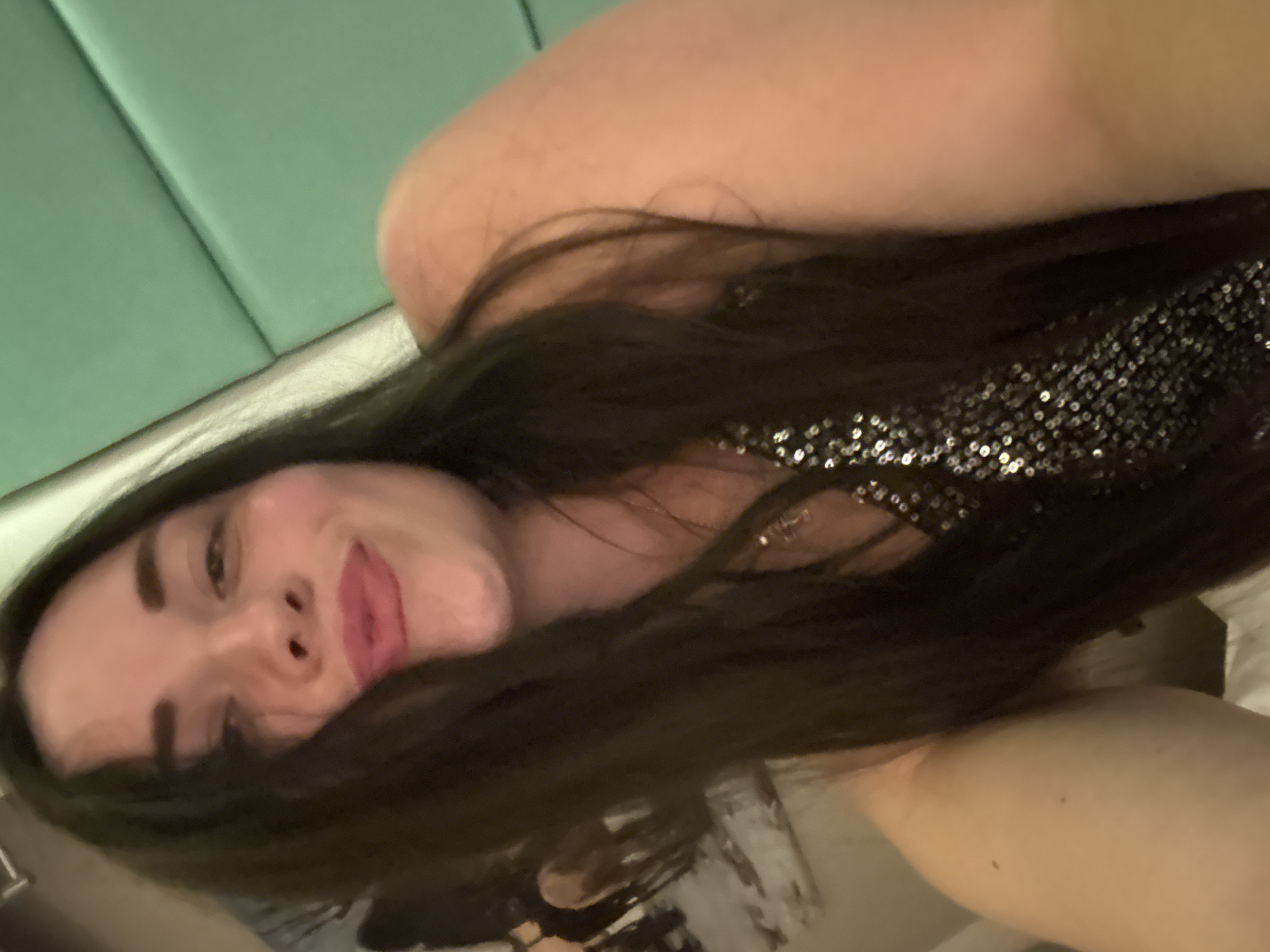A medium shot shows a woman with dark hair and light skin looking towards the camera. Her lips are parted in a pout, and she is wearing a dark, sequined dress. The background is out of focus, but appears to be a room with green, upholstered seating and a painting on the wall. There are no specific details in the image that would indicate a location in Al Hadaba, Egypt.