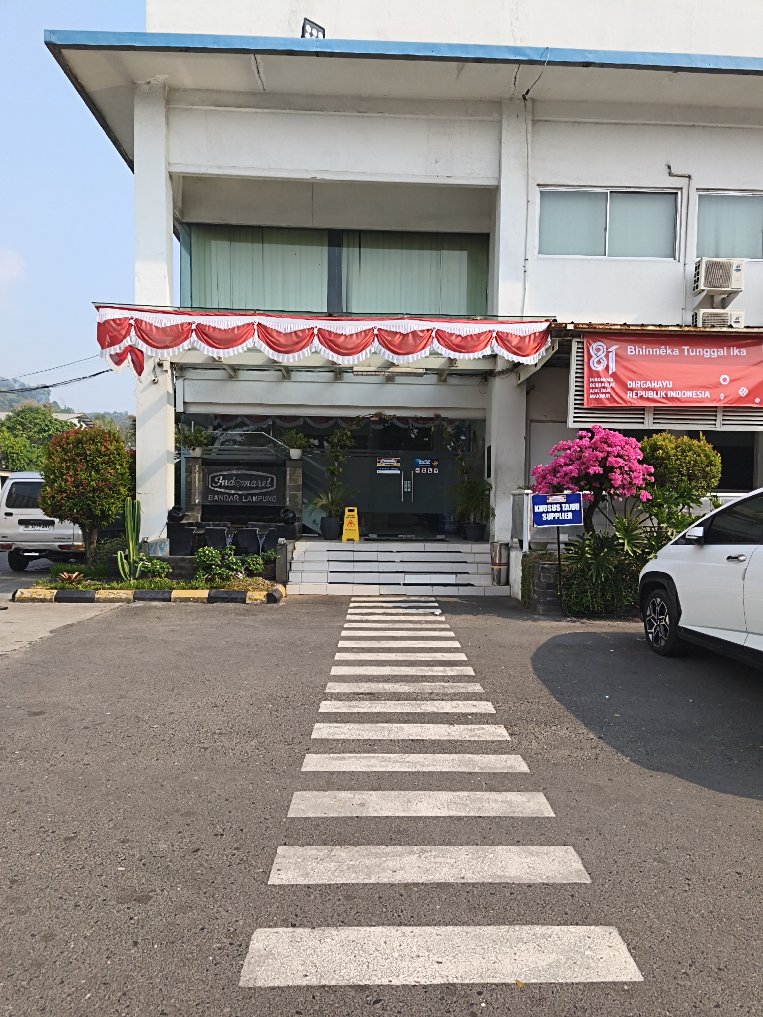 The image shows the exterior of an Indomaret convenience store located in Bandar Lampung, Indonesia. The building is a white, multi-story structure with a blue roofline. Red and white bunting is draped across the entrance. In front of the store is a pedestrian crossing painted on the asphalt. To the left, a silver van is parked. A decorative bush and a cactus are planted near the entrance. To the right, a white car is parked, and a pink flowering bush is in front of the building. A blue sign with white text reads "KHUSUS TAMU SUPPLIER," indicating a designated area for guest suppliers. The overall environment suggests a bright, sunny day.