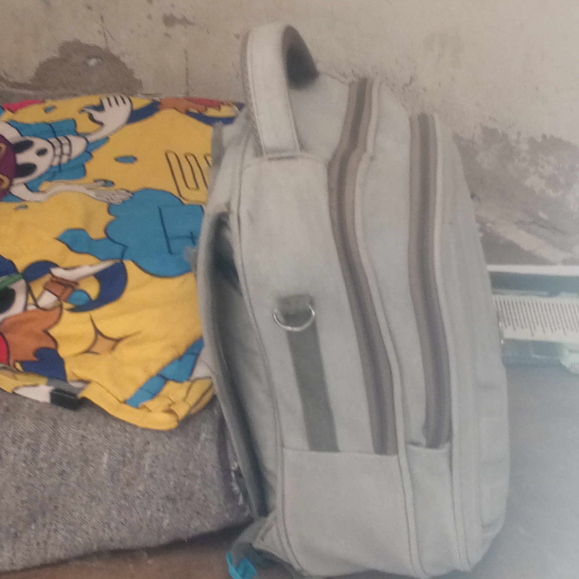 The image depicts a close-up view of a light gray backpack with multiple zippers and a fabric strap. The backpack is positioned vertically, leaning against a wall. To the left of the backpack, a folded piece of fabric with a colorful cartoon print is visible. The print features a character with a skeleton-like design on a yellow background with blue and red accents. The wall behind the objects appears to be textured and light-colored, with some scuff marks and possible water stains. The lower portion of the image shows a textured, gray surface, possibly a rug or a piece of furniture. The overall lighting is dim, with a slight glare on the right side of the backpack. There is no visible indication of the location in the city of Zinna, Nigeria.