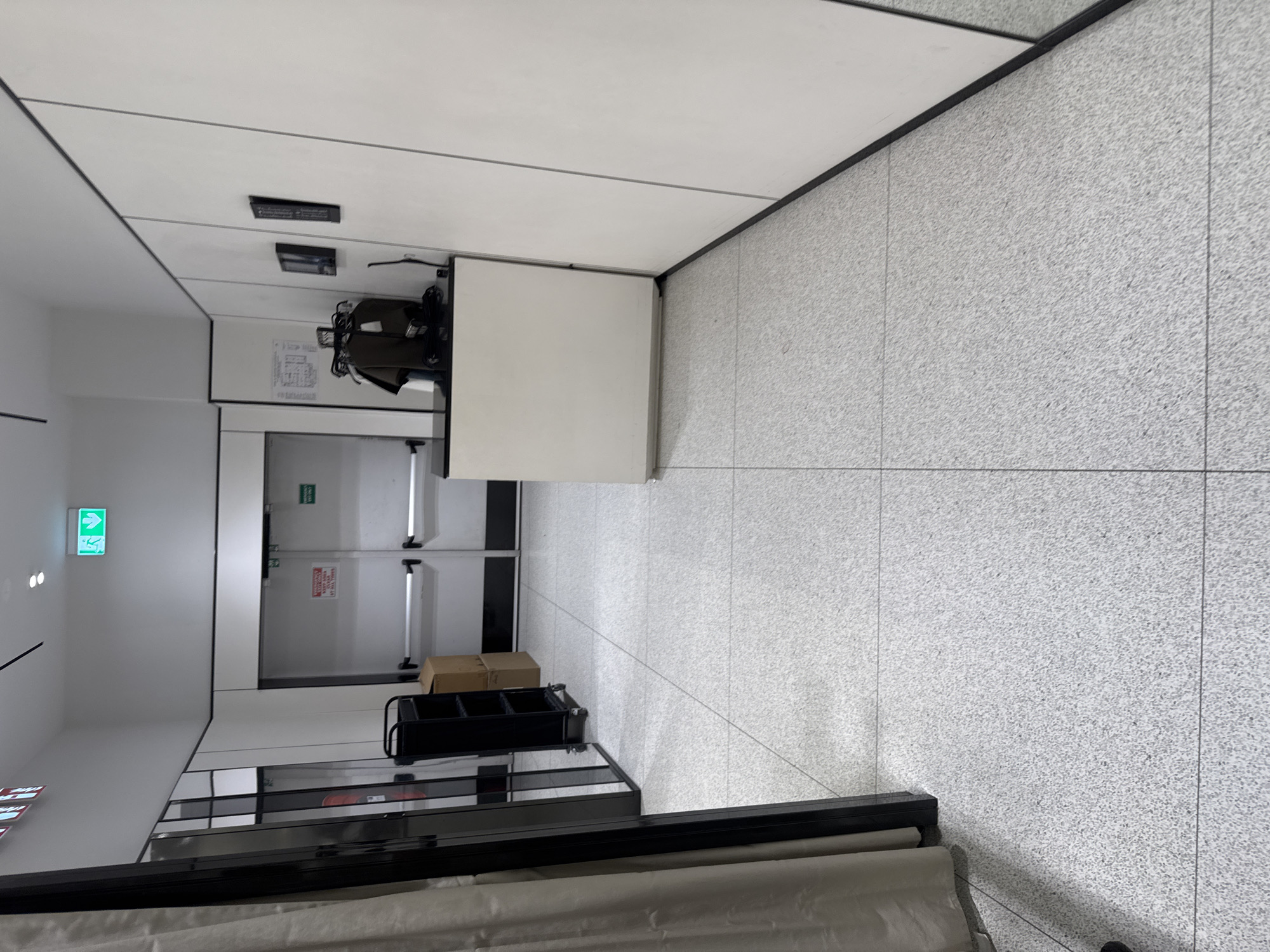 A modern, brightly lit indoor corridor is depicted, appearing to be in a commercial or public building in Plovdiv, Bulgaria. The perspective looks down the length of the corridor, which is empty of people.
The floor is covered in light grey, speckled tiles with visible grout lines. The ceiling features white panels separated by thin black lines, with several rectangular black light fixtures or vents. The walls are predominantly plain white.
On the left wall, an illuminated green exit sign is clearly visible, displaying a white running figure and an arrow pointing to the right. Below it, two small circular lights are present.
Further down the corridor, a set of double doors, possibly emergency exits, are at the end. These doors have silver push bars and a small, unreadable green sign above them. A red and white warning sign is partially visible on the left door, but its text is indistinct.
On the right side of the corridor, a white, rectangular counter or cabinet stands against the wall. Above it, a black object resembling a security camera or sensor is mounted. A diagram, possibly an evacuation plan, is affixed to the wall above the counter. Next to the counter, a black utility cart with multiple compartments holds a brown cardboard box.
The immediate foreground is partially obscured by a cream-colored fabric or rolled material. The scene suggests a clean, functional environment, likely during daytime given the bright artificial lighting, though the exact time is indeterminate. No specific activity is occurring.