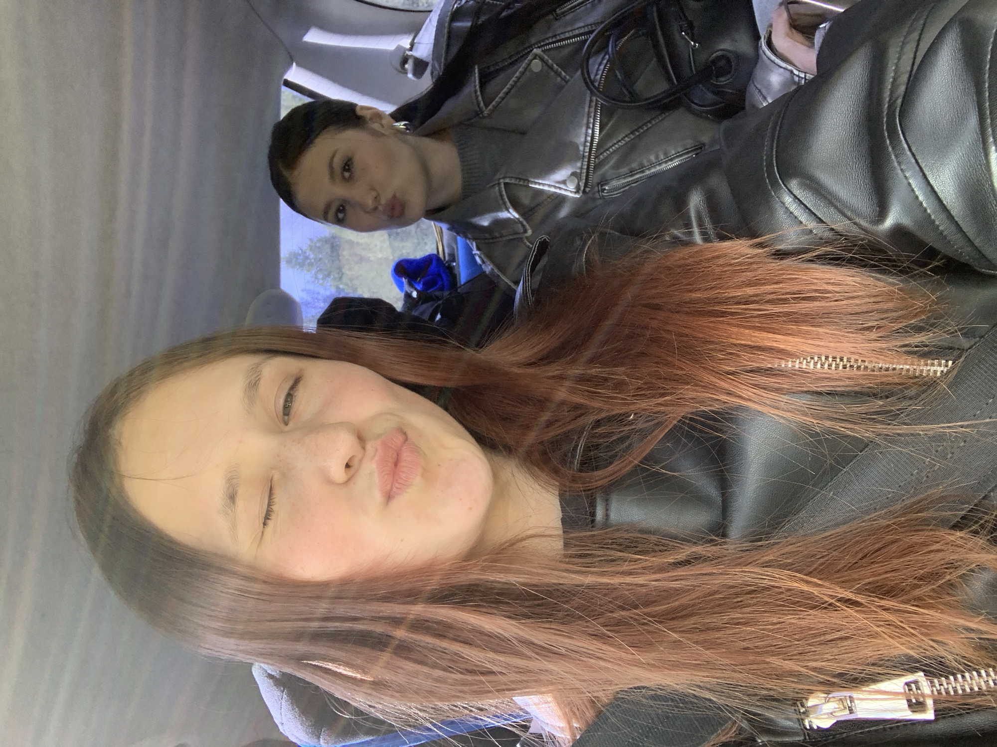 This media file captures two young individuals inside a vehicle, likely a car, in Chaikovsky, Russia, in what appears to be a selfie or casual photograph. The image is composed with one person in the foreground and another behind them.
The person in the foreground has long, reddish-brown hair, is wearing a black leather-like jacket, and is playfully winking with one eye while making a "kiss" face. Their expression conveys a lighthearted and casual mood.
Behind them and slightly to the right, a second person is visible, with dark hair pulled back. This individual is also making a "kiss" face and is dressed in a grey textured top beneath a grey or metallic-toned leather-like jacket, which features prominent zippers and snap buttons. A black bag or strap is partially visible near the second person.
The setting is the interior of a car, with parts of the ceiling and seating visible. Blurred outdoor elements, possibly trees, can be seen through a window in the background, indicating it is daytime. The overall emotion conveyed by both subjects is one of playfulness and ease. No text is visible within the image.