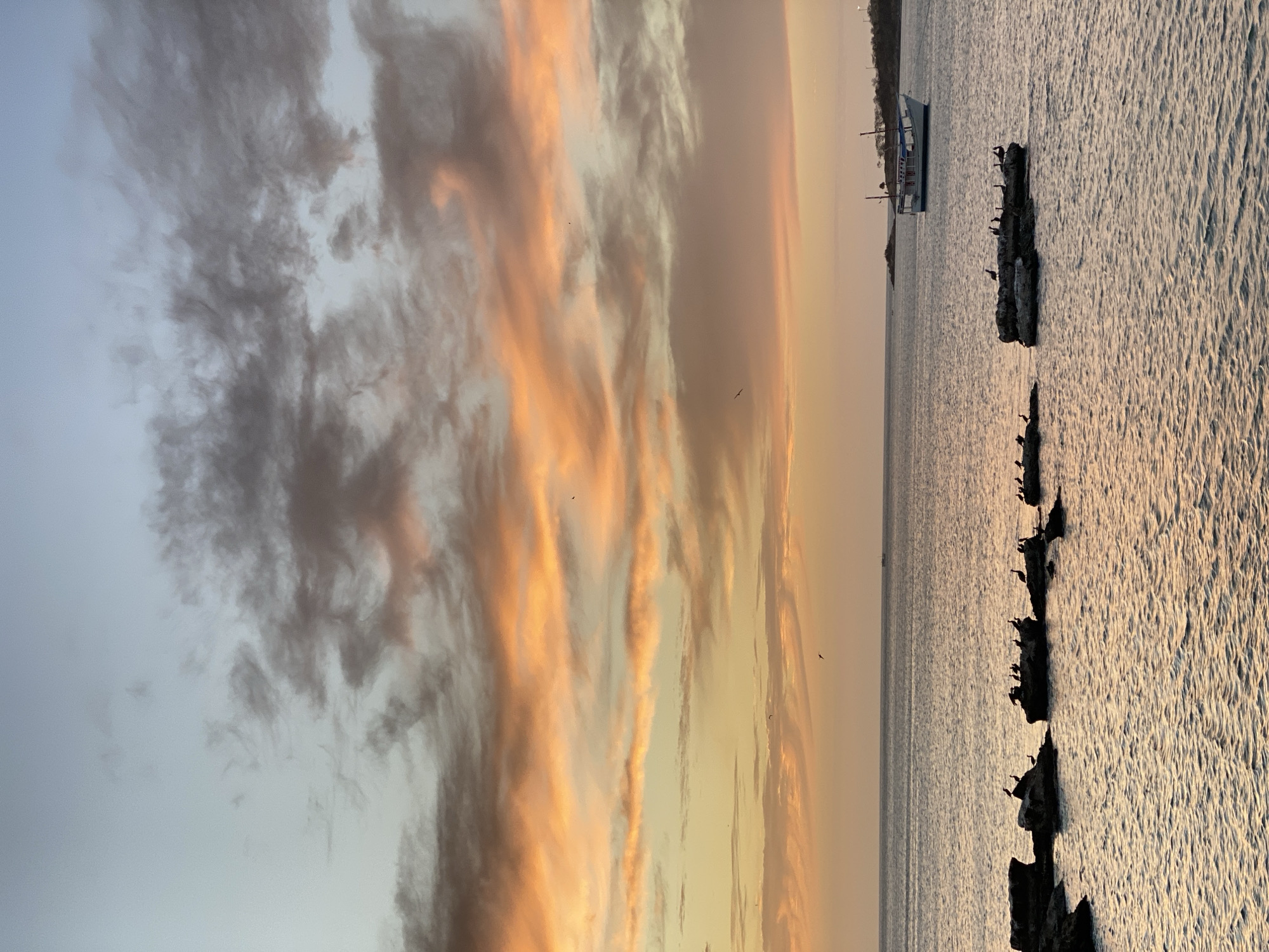 Here's a description of the image:
**Environment:**
The image depicts a sunset over the ocean, likely taken near the coastal city of Juan Griego, Venezuela. The sky is filled with clouds, with warm hues of orange and pink dominating near the horizon, transitioning to grays and blues towards the upper left of the frame. The water surface is slightly rippled, reflecting the sunlight.
**Objects/Subjects:**
* Several dark rocks or breakwaters extend into the water, and are populated by what appear to be birds, likely cormorants, perched on them.
* A boat is visible in the distance on the water.
**Actions/Interactions:**
* Birds are perched on the rocks.
* The sun is setting, casting light across the water and clouds.