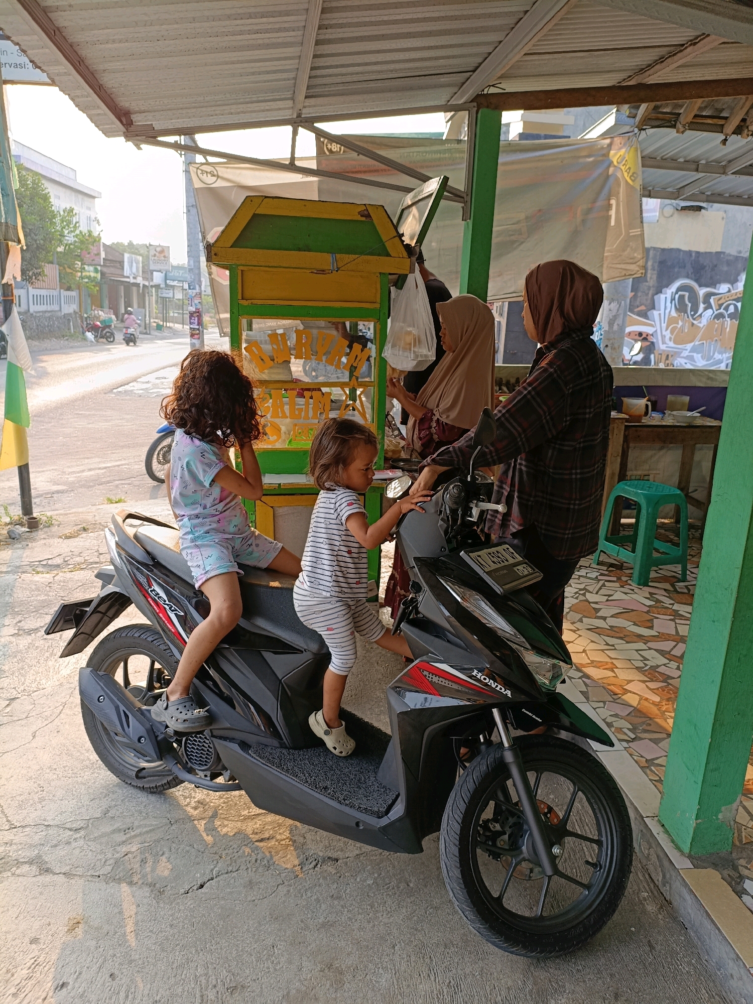 Two young girls and two adult women are in front of a food stall in Maguwoharjo, Indonesia. The girls, one older with curly hair and the other younger with straight hair, are sitting on a black Honda motorcycle. The older girl wears a multi-colored pajama set, and the younger girl wears a striped shirt and pants. One woman, wearing a brown hijab and a plaid shirt, stands next to the motorcycle, appearing to interact with the younger girl. Another woman, also in a hijab, is partially visible behind the food stall. The food stall itself is yellow and green, with signage that is partially obscured. The environment appears to be an outdoor setting with a paved ground and a building in the background. The sun is casting shadows, indicating it is likely late afternoon.