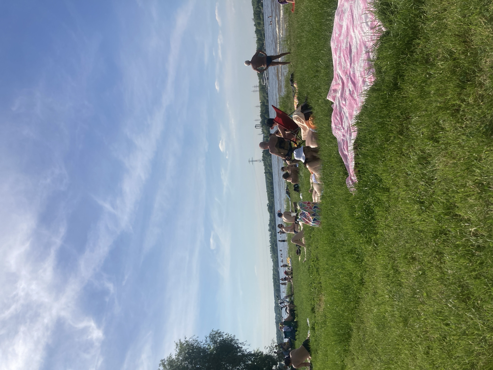 The image depicts a sunny leisure scene on a grassy bank next to a body of water in Kapustino, Russia. A clear blue sky with sparse, wispy white clouds covers the upper portion of the frame. The mid-ground features a wide expanse of water with ripples, suggesting a light breeze, and several individuals are visible swimming or wading further out. The far bank of the water body is lined with green trees and distinct industrial power line structures, including two tall pylons.
In the foreground and mid-ground, numerous people are present, mostly adults, engaged in various relaxed activities on the green grass. Many are seen lying on towels or directly on the grass, sunbathing, dressed predominantly in swimwear such as bikinis and swim trunks. Specific individuals include:
- A person in the bottom right foreground is lying down, with a large pink and white patterned towel spread beneath them.
- To the left of the pink towel, another person is lying prone, wearing a white bucket hat, with a multi-colored geometric patterned beach bag nearby.
- Further back, several individuals are seated in portable folding chairs, one prominently red and another dark blue, near a parked bicycle.
- On the right side, an older man with white hair, wearing swim trunks, stands upright at the water's edge with his hands on his hips, facing the water.
- Other individuals are scattered across the grassy area, relaxing in various positions. The overall atmosphere is one of casual recreation under bright daylight.
