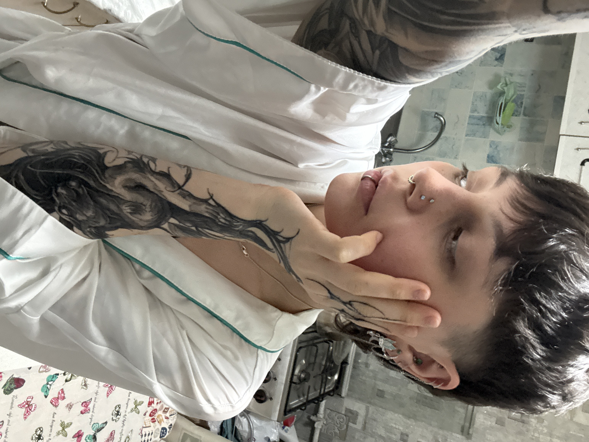 A person with tattoos is posing indoors in what appears to be a kitchen setting. The person is wearing a white robe with a teal trim. Their face is visible, showing several piercings, including a nose ring and a double piercing on the bridge of their nose. They have short, dark, wavy hair. The person's left arm is heavily tattooed, and they are touching their face with their left hand. Part of their upper torso is also visible, revealing more tattoos. The background suggests a kitchen with a stove and sink. There's also a table with a butterfly-themed tablecloth. Based on the architectural and interior design elements visible, there is no way to identify the location in the city of Saint Petersburg, Russia.