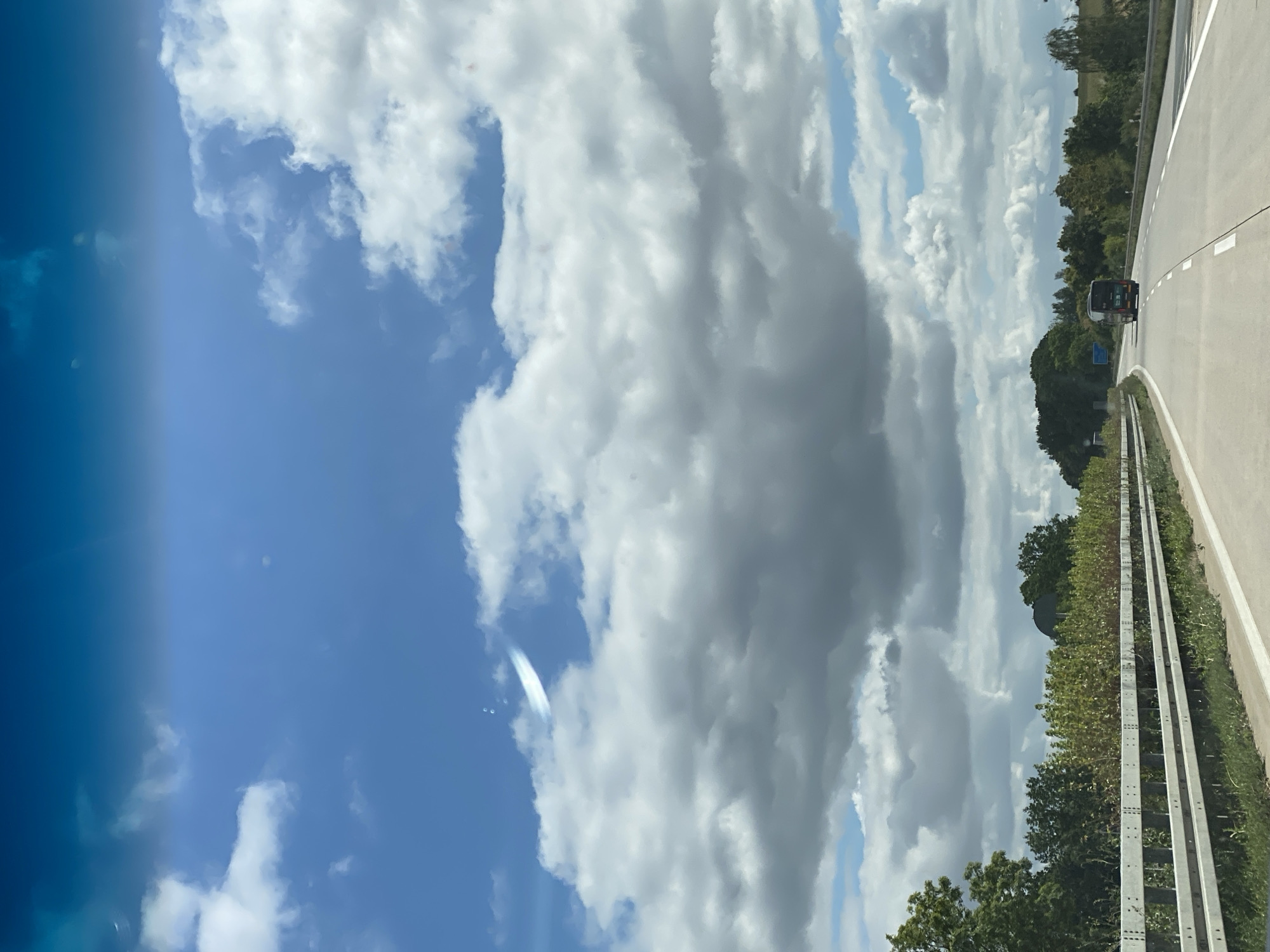A highway view shows a modern highway with multiple lanes, bordered by a metal guardrail on the left and lush green vegetation on the right. A dark-colored bus is visible in the distance, traveling on the highway. The sky above is a vibrant blue, dotted with fluffy white clouds, with a wispy contrail visible higher up. The scene appears to be a daytime shot taken from a moving vehicle, likely a passenger car, traveling along the highway in or near Nossen, Germany. The landscape suggests a rural or suburban setting outside of the city center.