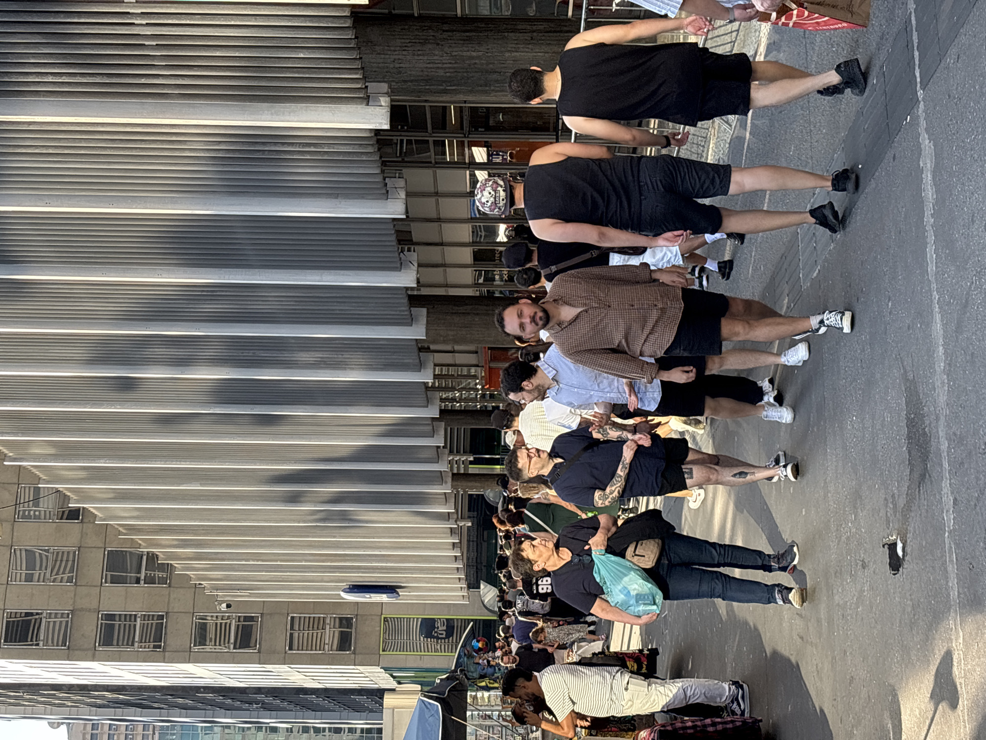 The image shows a crowd of people walking along a street in São Paulo, Brazil. The majority of people are dressed casually, with a variety of skin tones and ages represented. A few individuals are closer to the camera and are walking away from the viewer. A building with a corrugated metal facade is visible on the left side of the frame. Other buildings and structures are visible in the background, as well as vendor booths. The street appears to be paved.