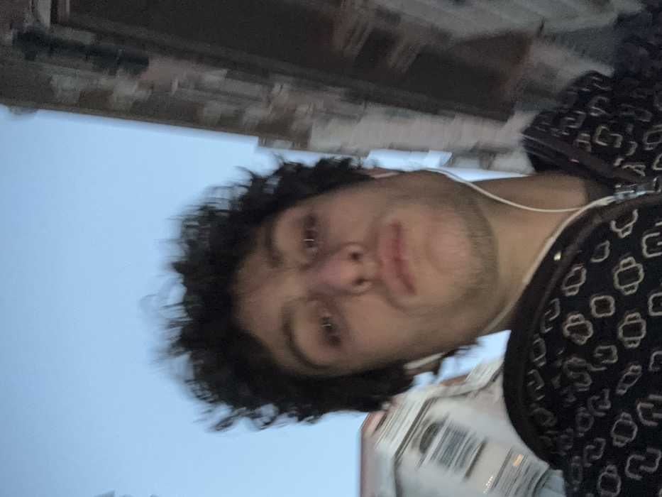 This image is a close-up, low-angle selfie of an adult male in an urban setting, consistent with the city of Vincennes, France. The male subject, approximately in his mid-20s, has dark, curly hair and light stubble on his jawline and chin. He is wearing a dark-colored, zippered jacket or hoodie with a repeating pattern of light-colored, stylized geometric shapes. White wired earbuds are inserted into both ears, with the wires visible extending downwards. His gaze is directed forward, directly at the camera, with a neutral facial expression. The sky above is a clear, light blue. In the blurred background, architectural elements are visible: a light-colored, multi-story building with a visible arched window is present on the upper left, while a darker, textured building facade is partially visible on the upper right. The background elements appear out of focus compared to the subject. The scene is illuminated by natural, even light.