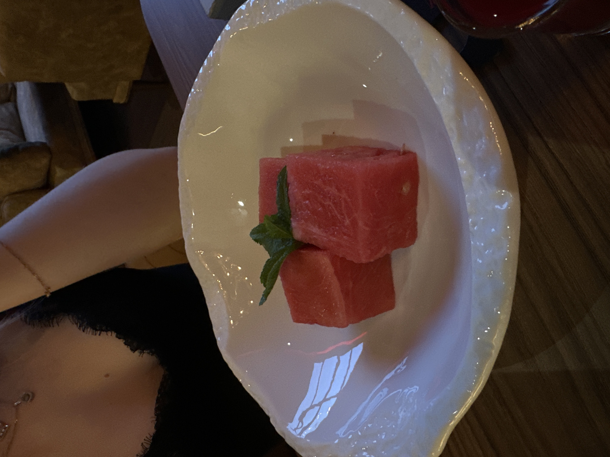 The image captures a close-up, slightly overhead view of a white, irregularly shaped bowl, possibly ceramic, containing a dessert. Inside the bowl are two cubed pieces of red watermelon, with one stacked on top of the other, and a small sprig of green mint resting between them. The bowl has a glossy surface, reflecting light from a window or light fixture.
To the left of the bowl, a person's bare arm, shoulder, and a portion of their upper chest/neck are visible, wearing a dark-colored top, possibly black, with a slightly textured edge near the neckline. A thin, golden chain or bracelet is visible on the person's wrist, and a small mole is present on their chest. In the upper right corner, part of a glass with a dark red liquid is visible.
The scene takes place indoors, likely at a table made of dark wood, suggested by the blurred background. Other blurry elements in the background hint at furniture, possibly a couch or chairs. The lighting appears to be ambient, suggesting an evening setting or a dimly lit interior. The image depicts a moment of enjoying a refreshing snack or dessert. No text is visible. The location context is Vladimir, Russia.