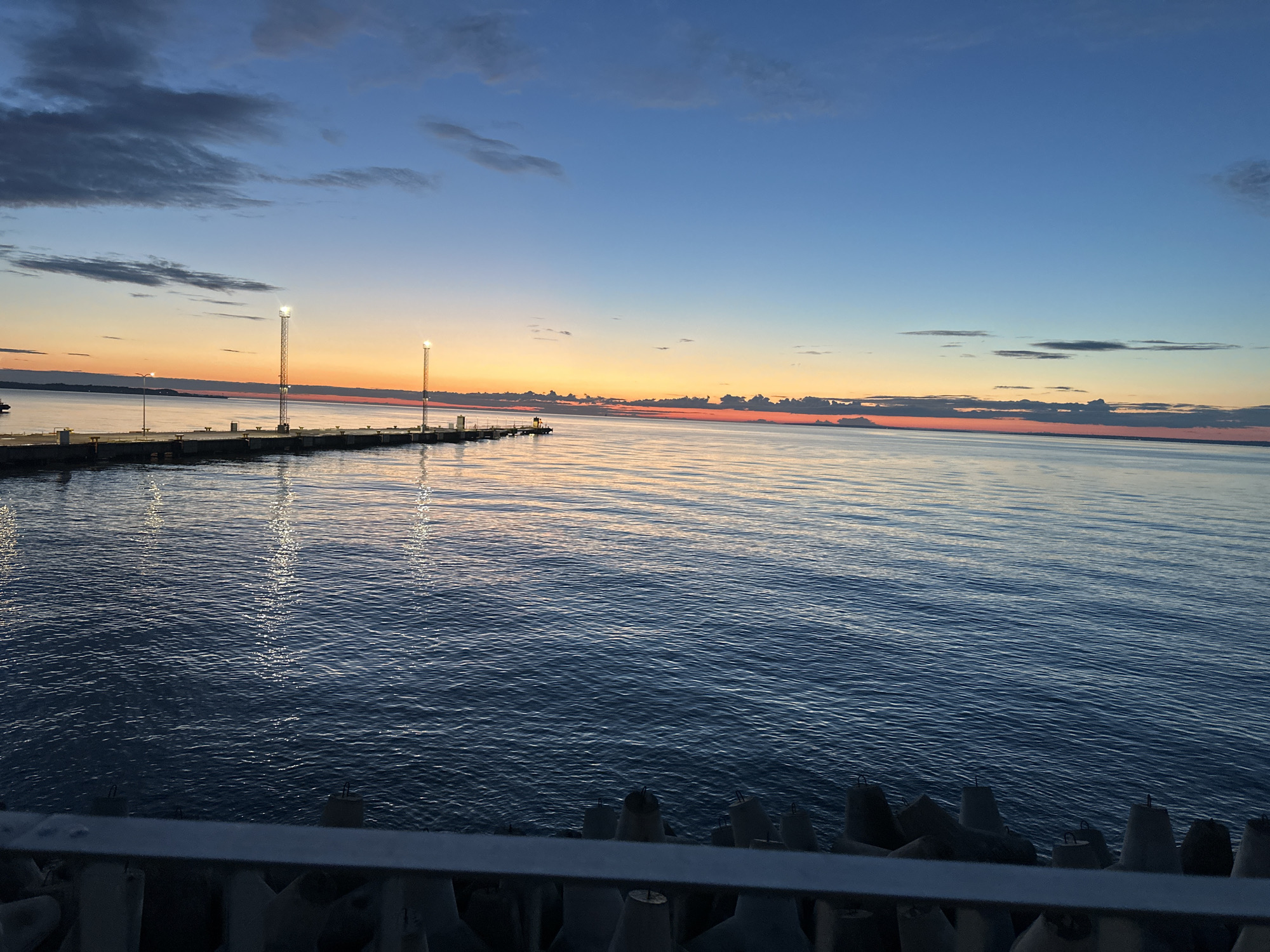 A wide-angle, eye-level shot captures the serene seascape of Tallinn, Estonia, during twilight. The sky transitions from a deep blue at the zenith to a warm gradient of orange and pink near the horizon, indicative of a sunset or sunrise. Wispy clouds dot the upper atmosphere. In the middle ground, a long, dark pier juts out into the calm, deep blue water, illuminated by several tall light poles, their reflections shimmering on the water's surface. The water exhibits gentle ripples, creating a textured expanse. In the foreground, a grey railing is visible, with the tips of several concrete tetrapods, commonly used for coastal protection, peeking out from beneath it. The overall ambiance is one of tranquility and natural beauty, with the man-made structures of the pier and harbor defenses harmonizing with the expansive maritime landscape.