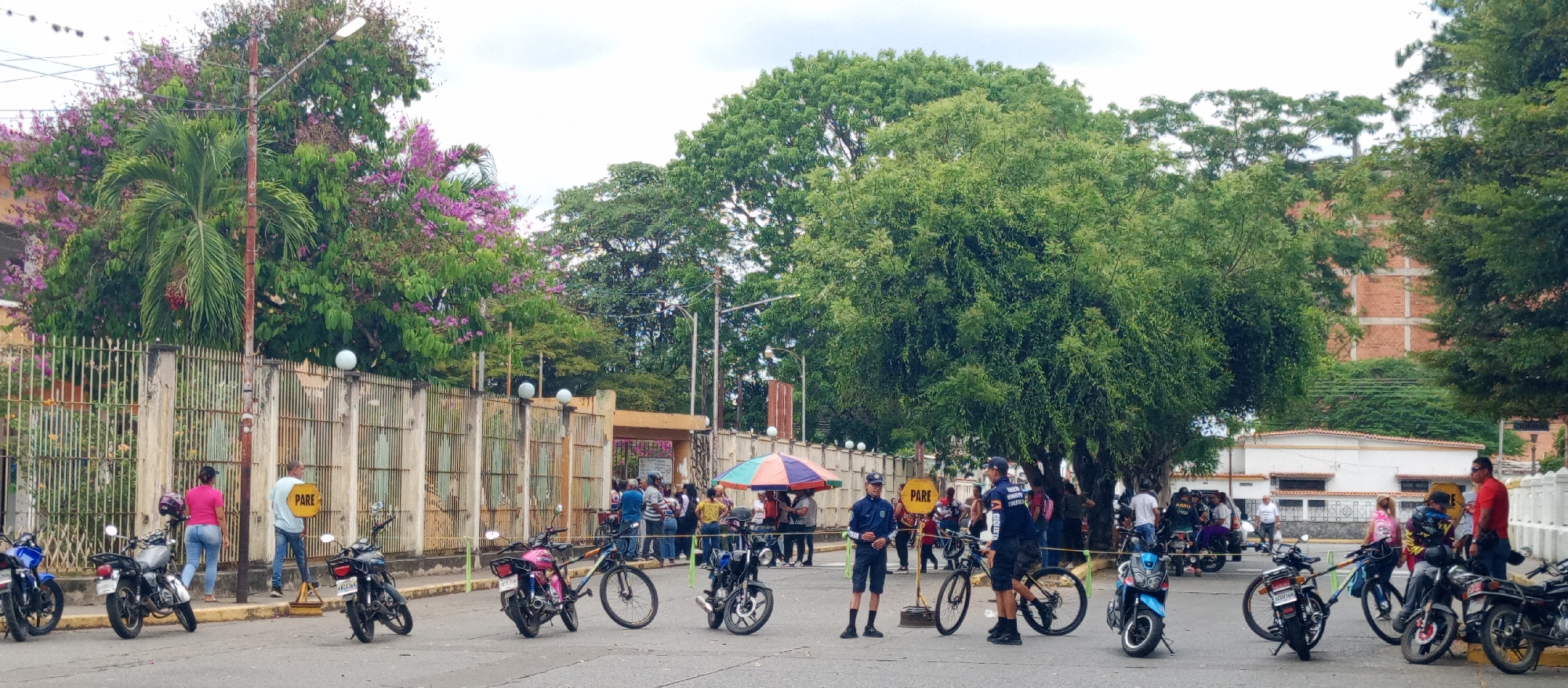 The image depicts a street scene in Tinaquillo, Venezuela, with a crowd of people gathered near a gated structure. Multiple motorcycles and bicycles are parked or positioned along the street. Several individuals appear to be law enforcement or security personnel, wearing uniforms and standing near the parked vehicles and a "PARE" (STOP) sign. Other people are standing around, talking, and observing. A large tree dominates the right side of the frame, while a flowering tree is seen on the left. A building with brickwork is visible in the background behind the trees.