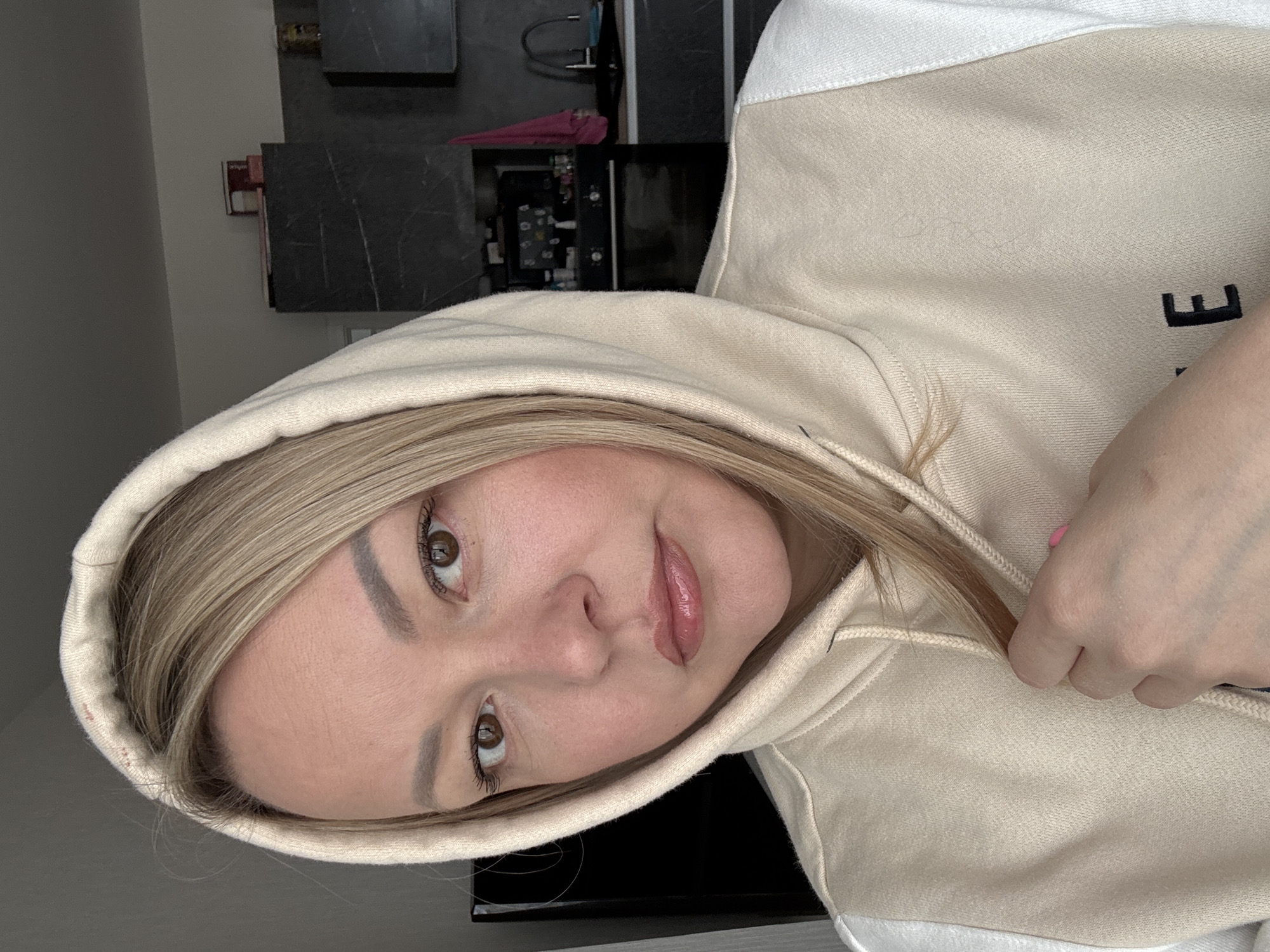A woman with light blonde hair, wearing a beige hoodie, is looking at the camera. Her face is slightly tilted. The background includes what appears to be a kitchen area with dark cabinets and appliances. The location is unknown, and there is no indication that the scene is in Izhevsk, Russia. The hoodie has lettering partially visible on the side. The woman's hand is partially visible, near the drawstring of the hoodie.