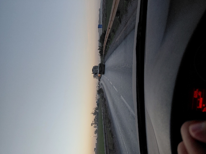 A driver's perspective reveals a road ahead under a bright sky, possibly during dawn or dusk. Vehicles are visible in the distance. The road appears wet, and a grass verge is present to the side. The car's dashboard and speed display are visible, with a hand partially obscuring the display. The scenery gives the impression of a highway or expressway journey.
