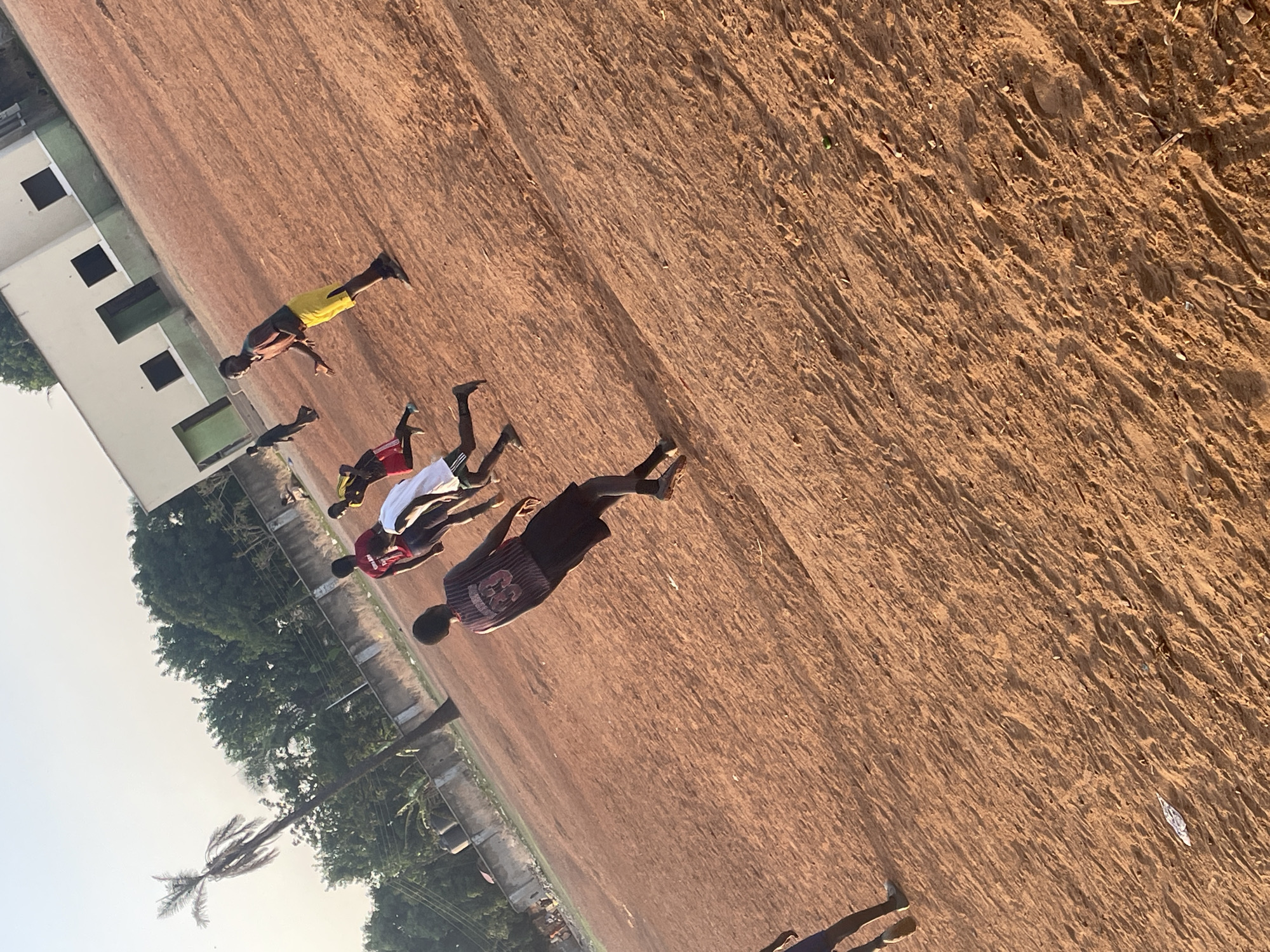 The image depicts a group of young males gathered on a large, reddish-brown dirt field, likely in Akwanga, Nigeria. The scene is bathed in warm, soft light, suggesting either early morning or late afternoon with a hazy sky.
At least six individuals are visible. They are dressed in casual athletic wear, including shorts and t-shirts or jerseys. One person prominently wears a dark jersey with the number "33" on the back, while another is in yellow shorts and a dark top, appearing to be in mid-stride or a dynamic pose. Another individual wears white shorts and a red top. Their poses suggest they are either engaging in a sports activity, possibly football (soccer), or are in a state of rest or preparation on the field.
In the background to the left, a light-colored, multi-story building with several dark, square windows stands, possibly a school or residential complex. Lush green trees and foliage flank the building and extend along the horizon, with a tall, slender palm tree distinctly visible on the far left. The ground itself is uneven, showing textures of packed earth and loose soil with some small scattered debris. The overall setting portrays an active outdoor community space.