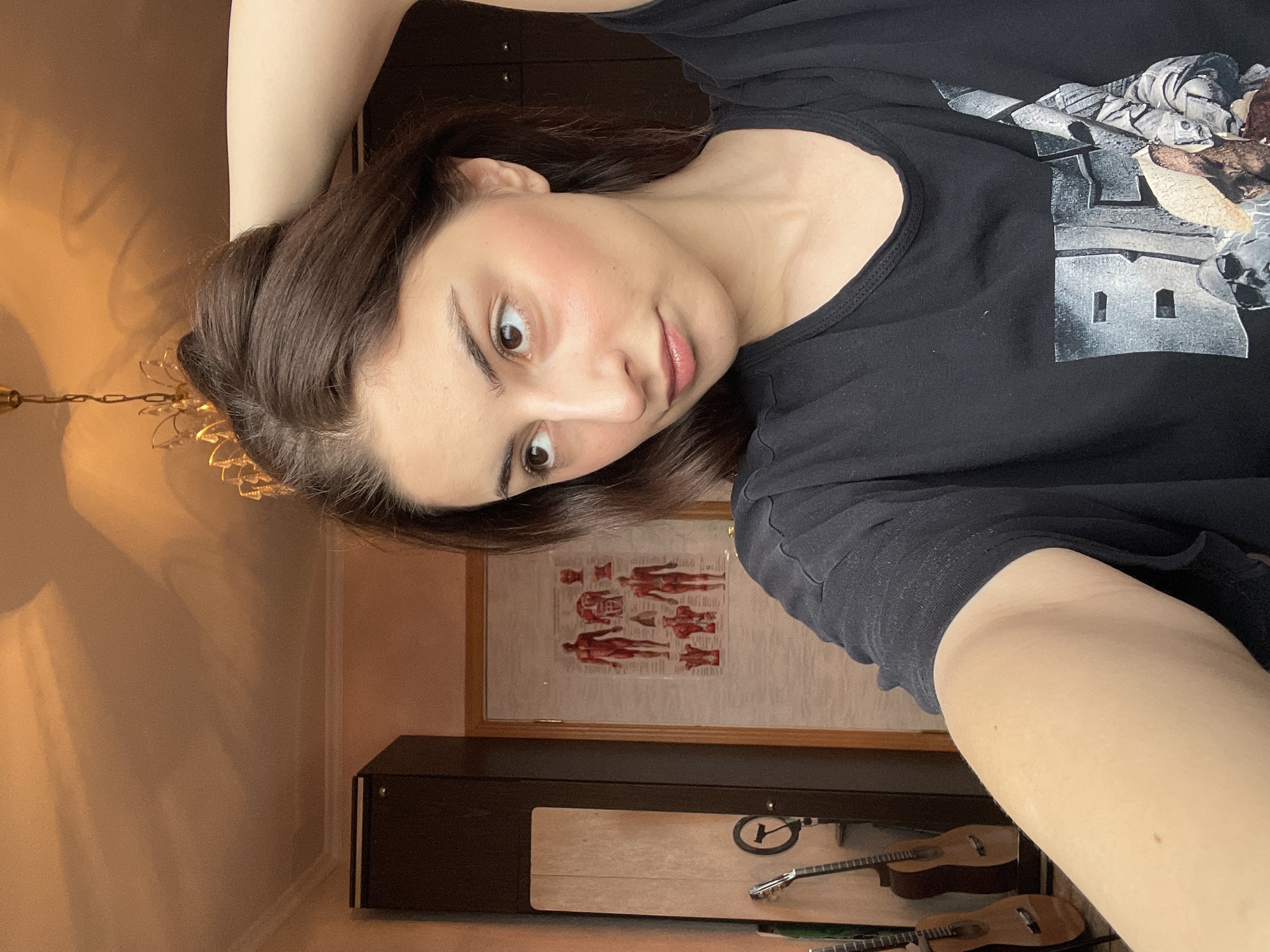 A person with dark hair is lying down, looking at the camera. The person is wearing a black t-shirt with a design on it. Behind the person is a wardrobe, a framed poster of human anatomy, and two guitars in their cases. There is also a chandelier visible on the wall. It is impossible to ascertain if the location is in Moscow, Russia.