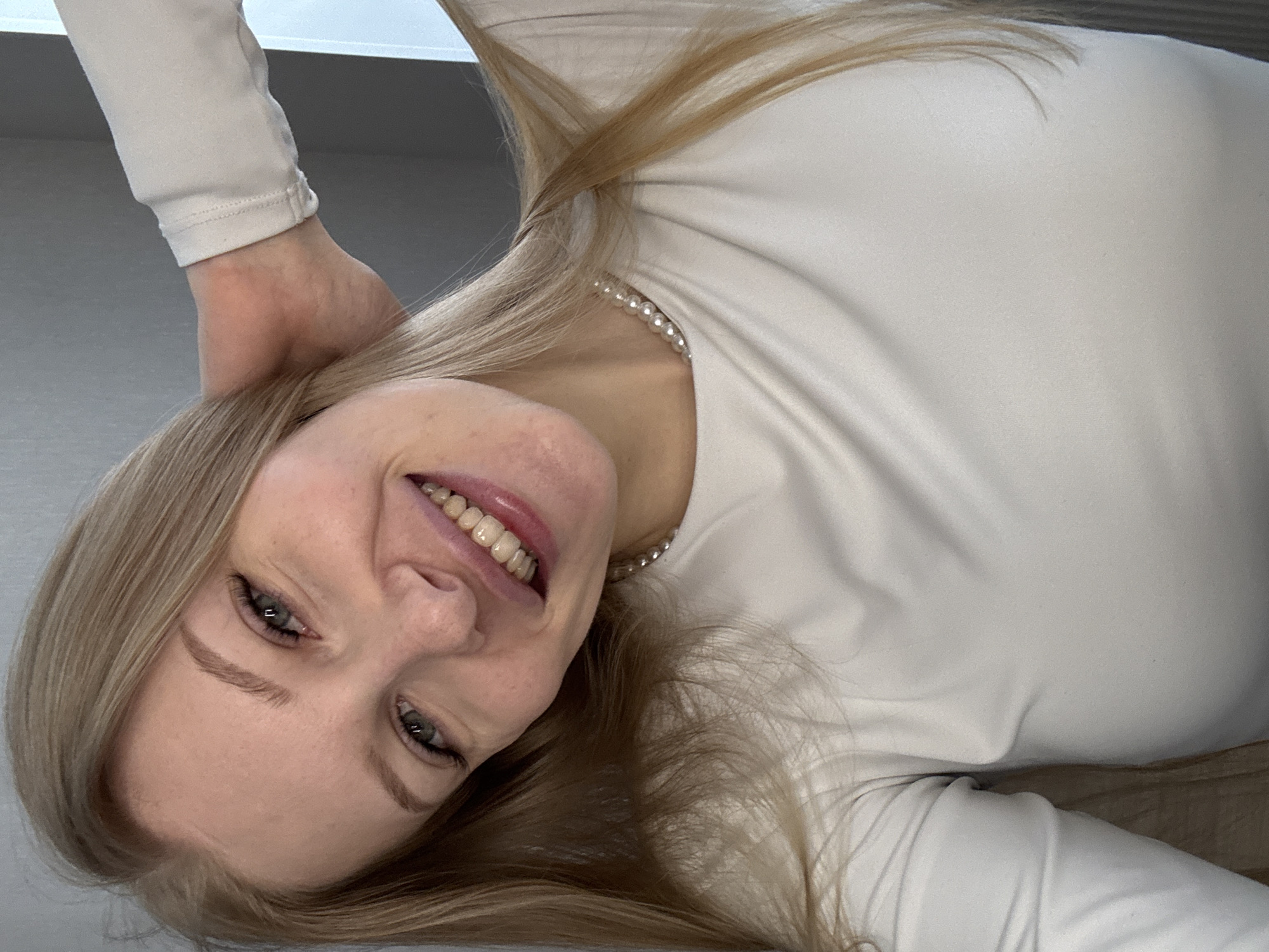 A cheerful young woman with long blonde hair is depicted in a close-up, overhead shot. Her face is inverted relative to a standard upright perspective, as if she is lying down and looking up at the camera. She is smiling broadly, revealing her teeth, and her eyes are open and engaging the viewer. Her blonde hair spreads out around her head. She is wearing a light-colored, possibly white or off-white, long-sleeved top or dress. A delicate pearl necklace is visible around her neck. Her left hand is seen resting near her head, blending with her hair. The background is indistinct and uniformly light-toned, suggesting an indoor setting with plain walls or a ceiling. The lighting is soft and even, without harsh shadows. No text is visible in the image.