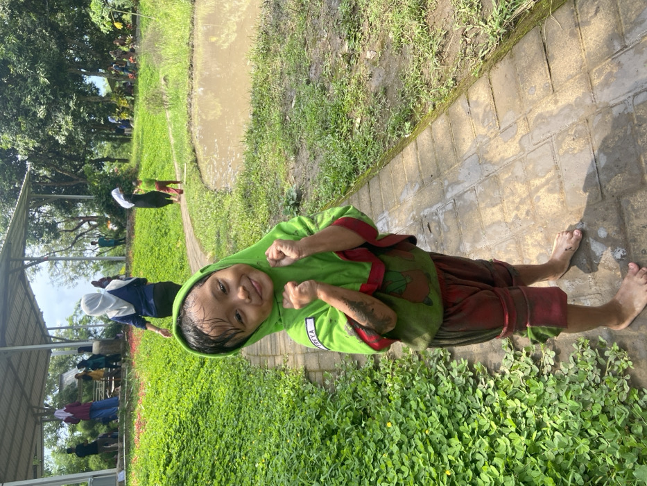 A young child with a dirty face and hands stands barefoot on a brick path, smiling and holding their fists up. The child wears a green hooded sweatshirt and reddish-brown shorts. Behind them is a grassy area with a muddy ditch or pond, where people are gathered under a long shelter. Trees are visible in the background. The scene appears to be outdoors, possibly in a rural or park setting.